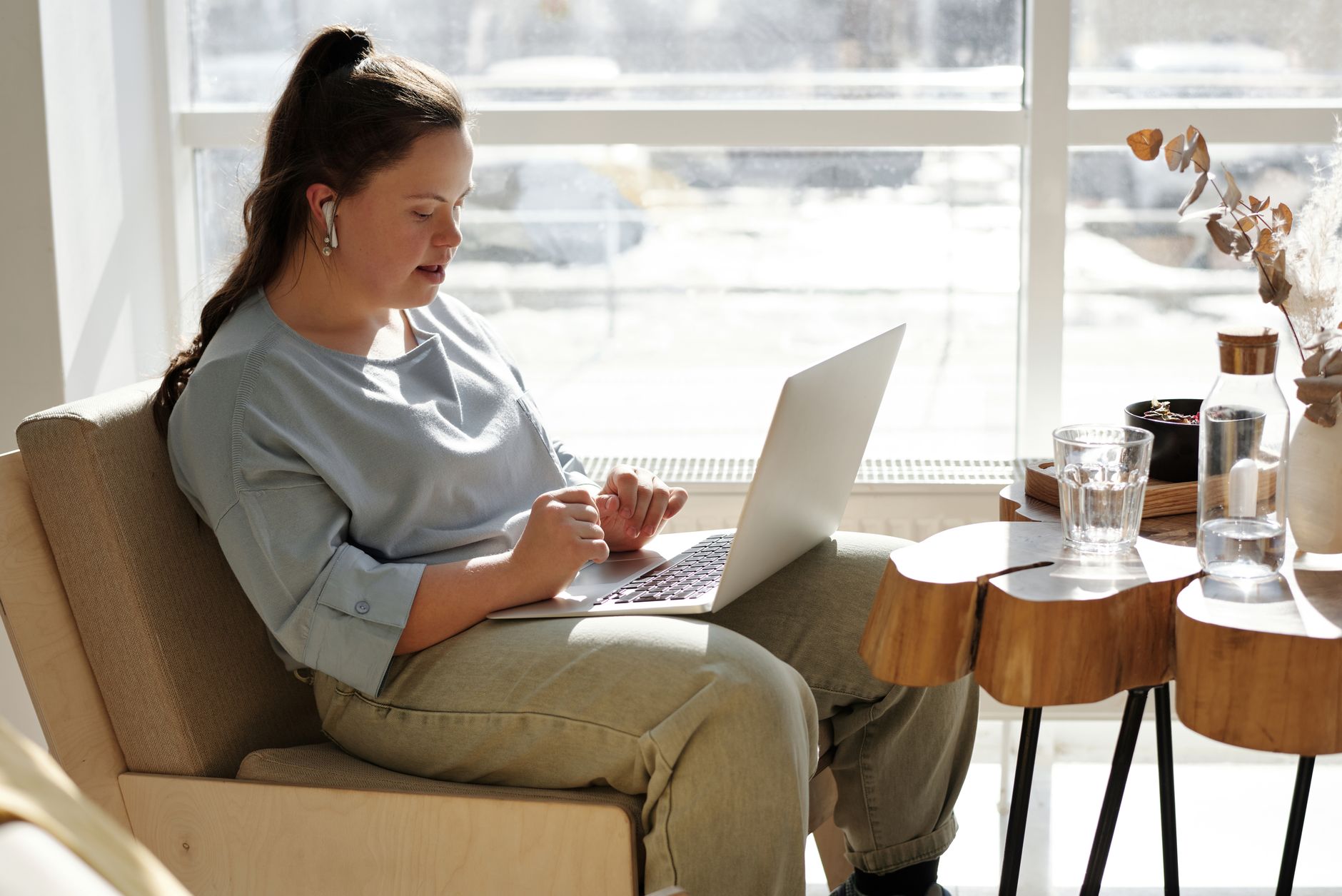 A young woman with Down syndrome sits in an armchair by a sunny window, wearing earbuds and typing on a laptop