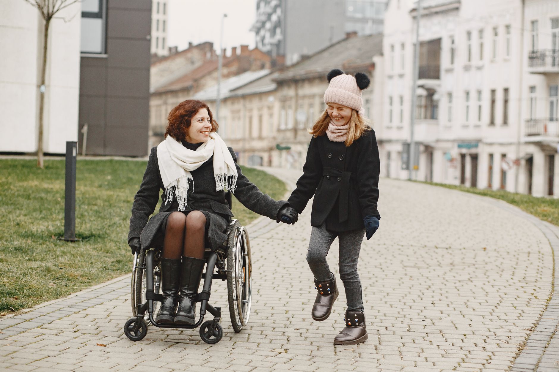 A smiling woman in a manual wheelchair holds hands with a laughing girl on a paved city path in winter coats