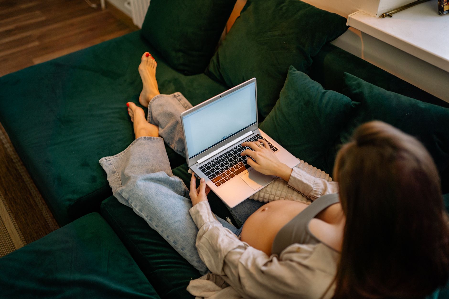 A pregnant woman sits on a green sofa typing on a laptop