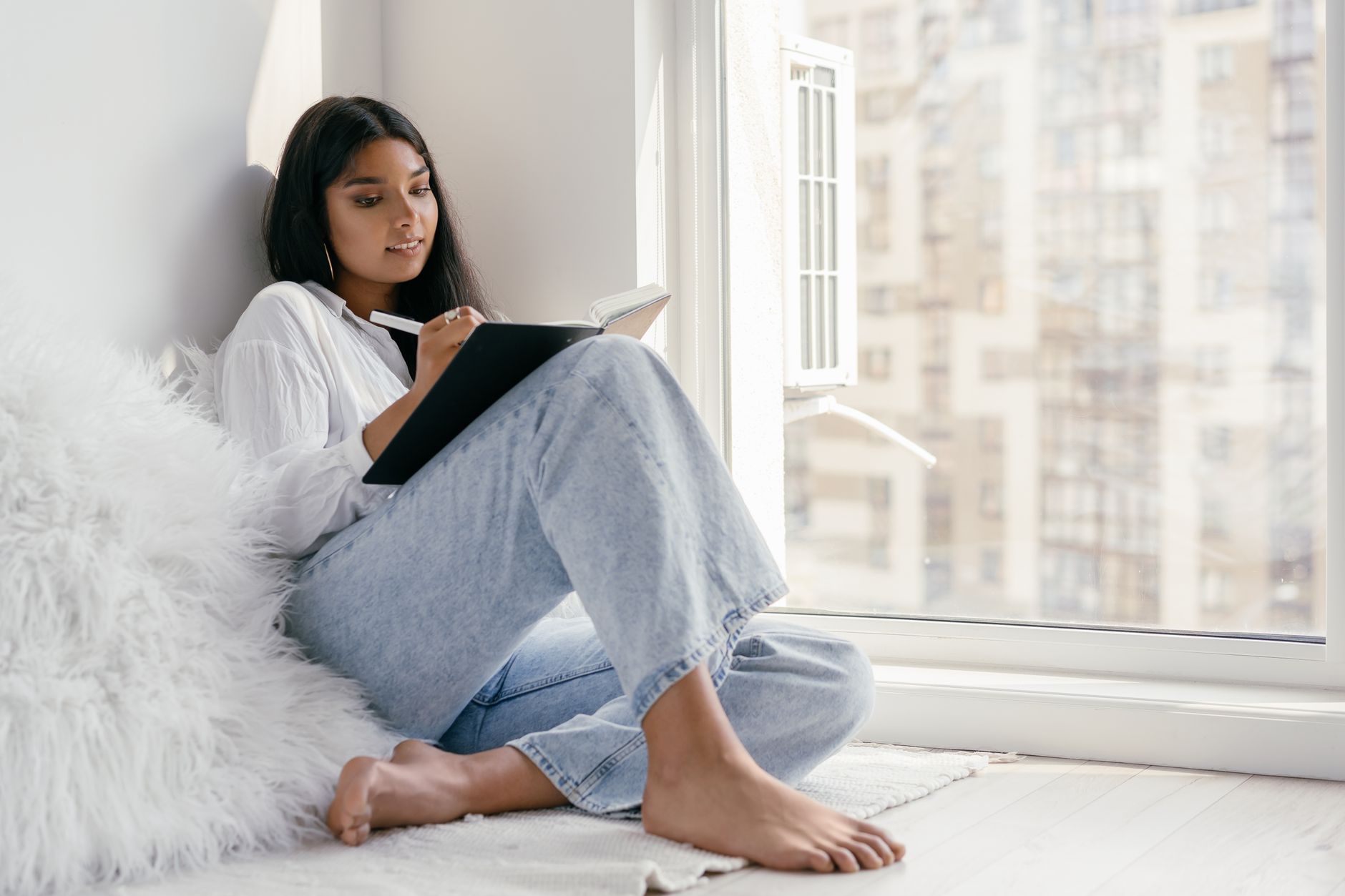A young woman in a white shirt and jeans sits on the floor by a sunny apartment window, writing in a notebook