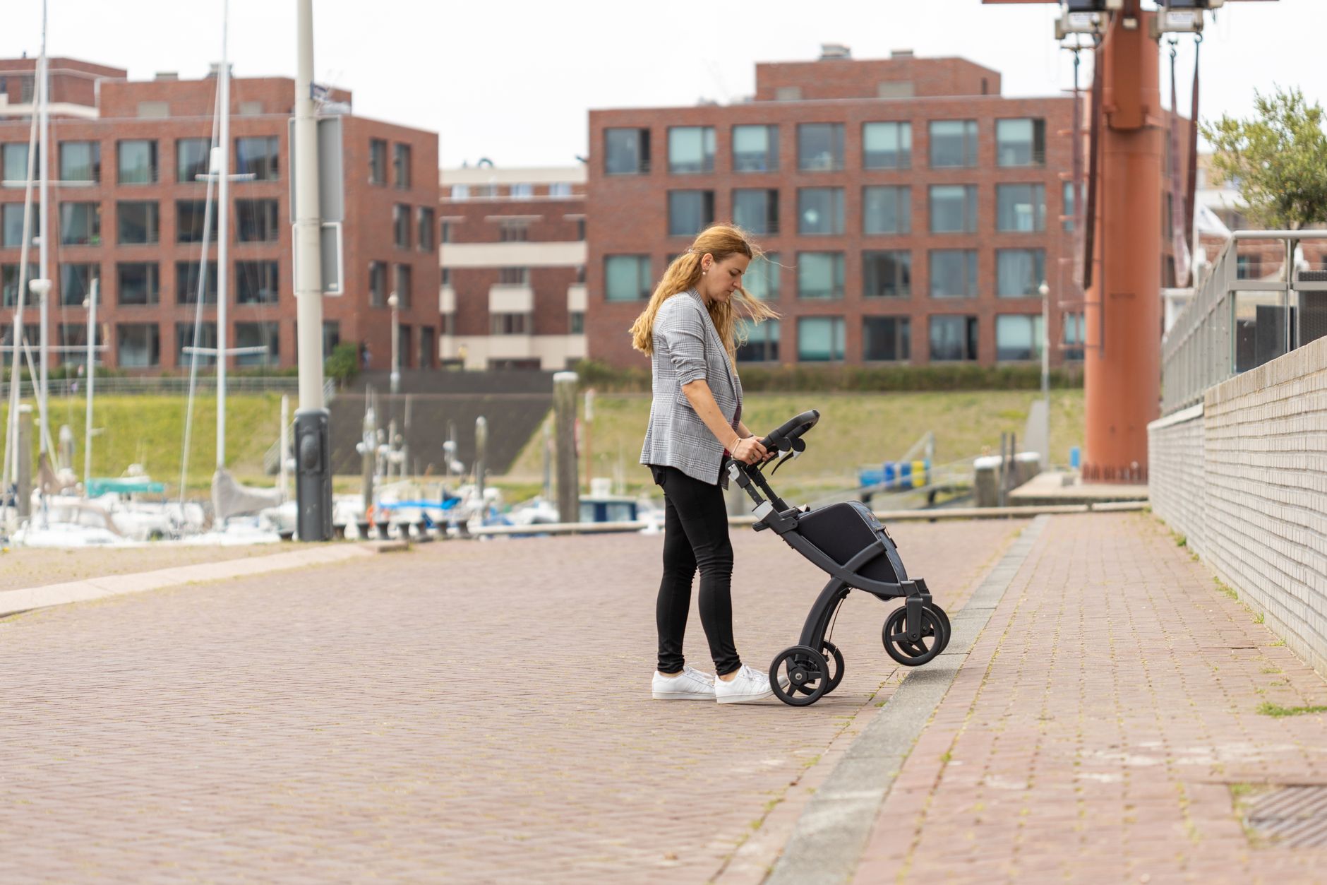 A woman in a gray blazer guides a rollator walker along a brick waterfront promenade beside a marina