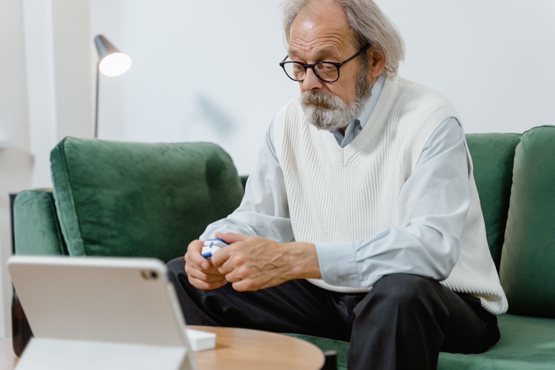 A gray-bearded older man in glasses and a white sweater vest checks a pulse oximeter on his finger while sitting on a green sofa