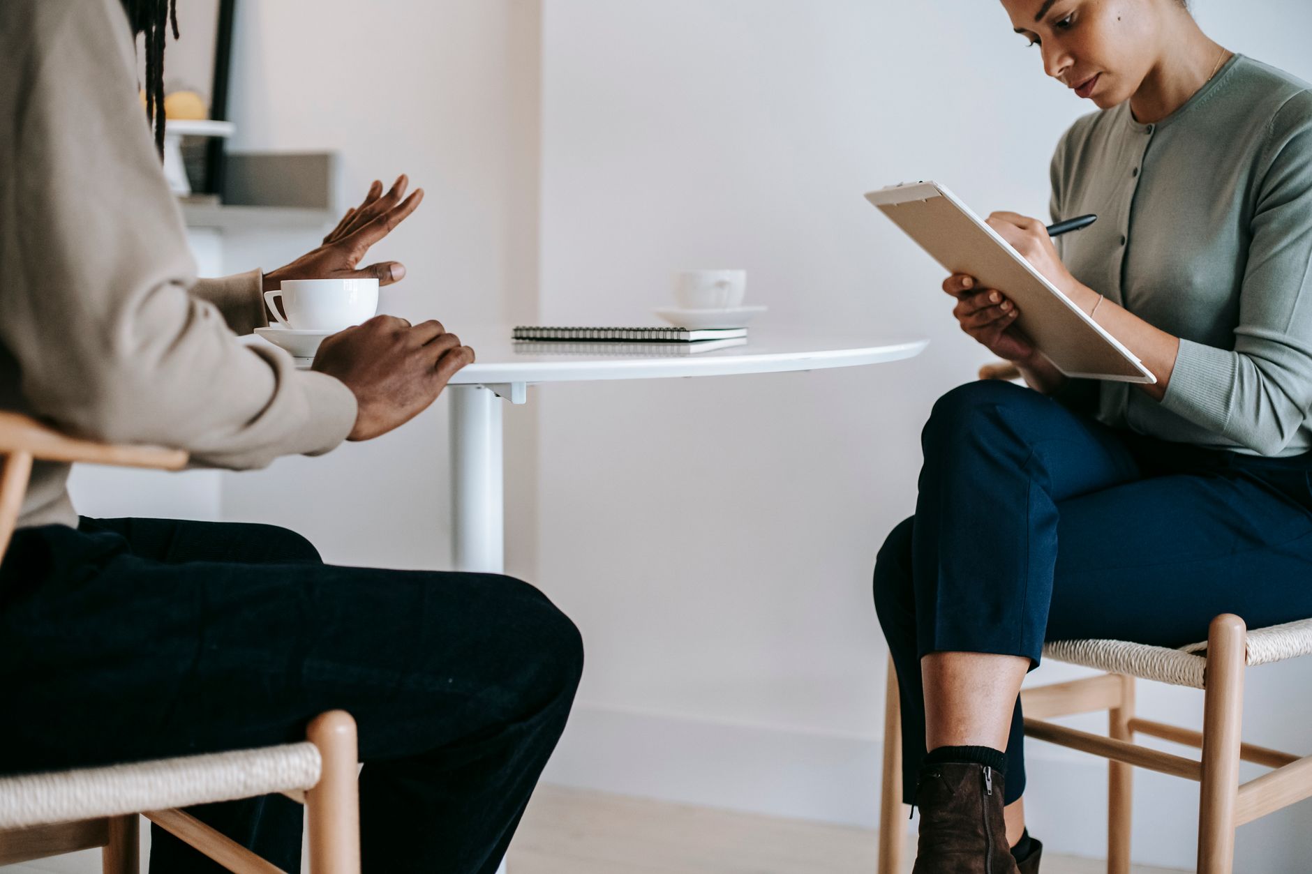 A counselor in a green cardigan writes on a clipboard while a man across a small white table talks and gestures