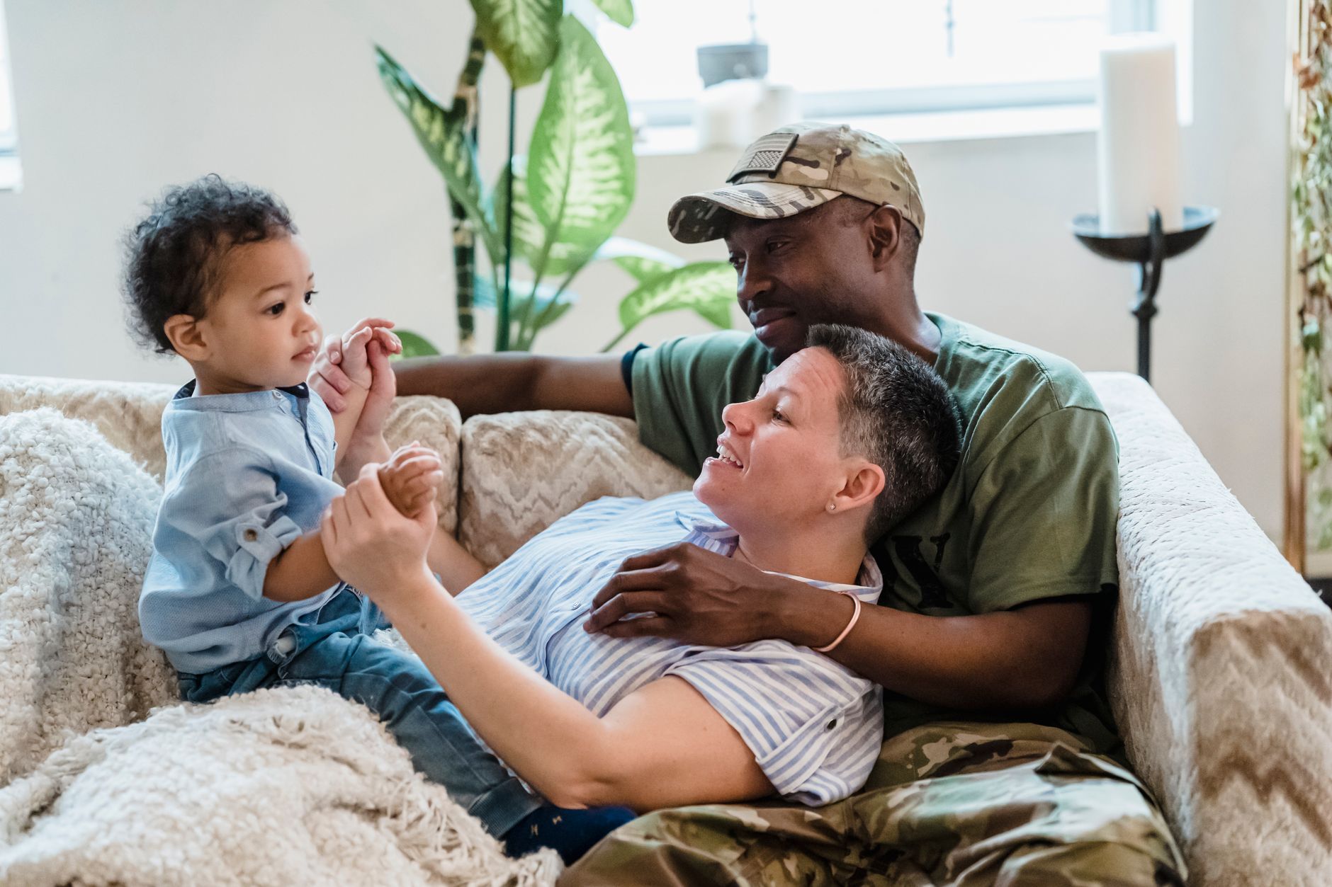 A man in a camouflage cap and green T-shirt smiles as his partner plays with their toddler on a living room sofa