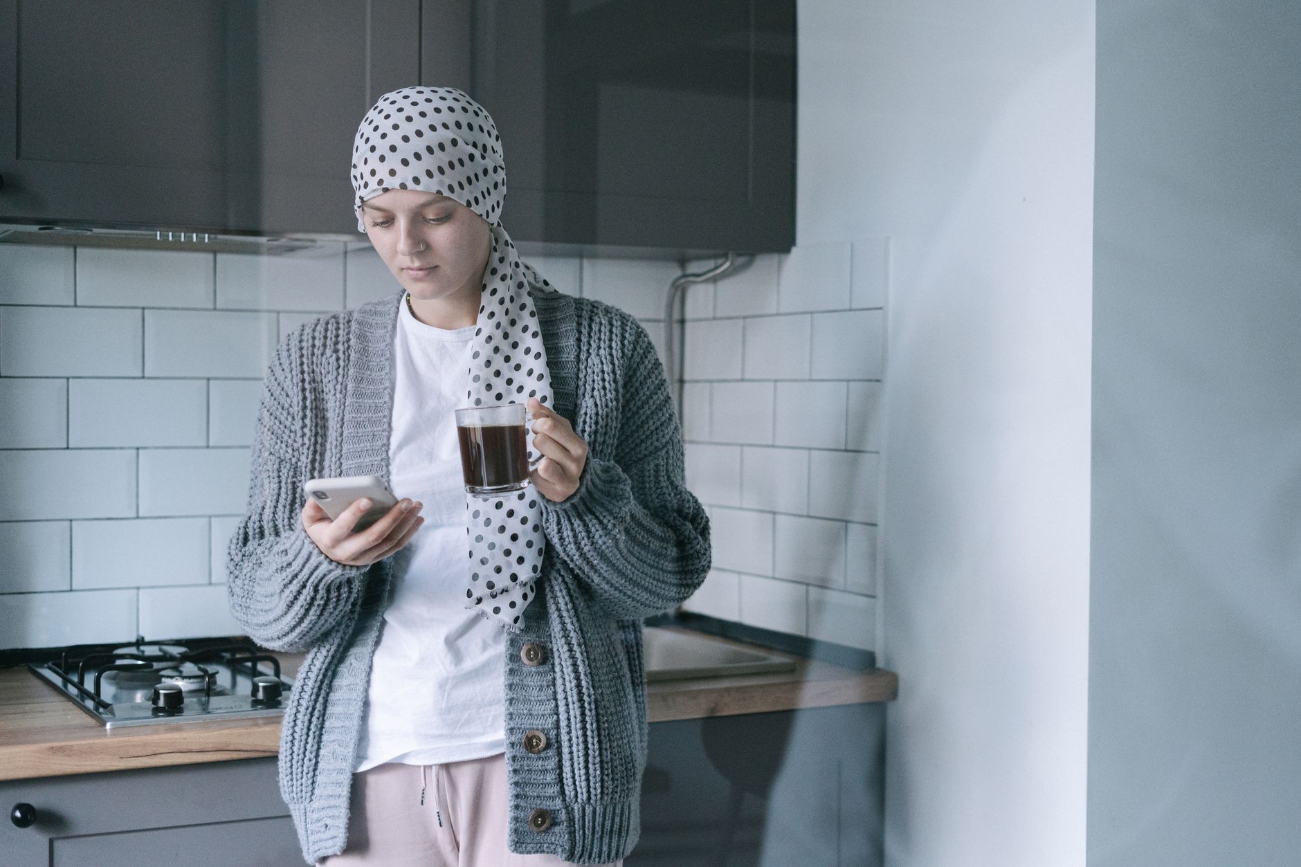 A woman wearing a polka-dot headscarf and gray cardigan reads her phone while holding a cup of tea in her kitchen