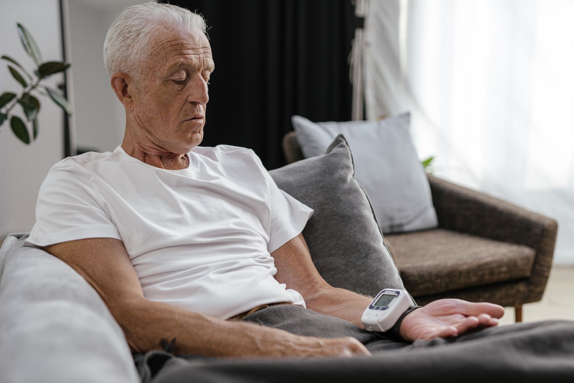 An older man in a white T-shirt sits on a sofa at home checking a wrist blood pressure monitor