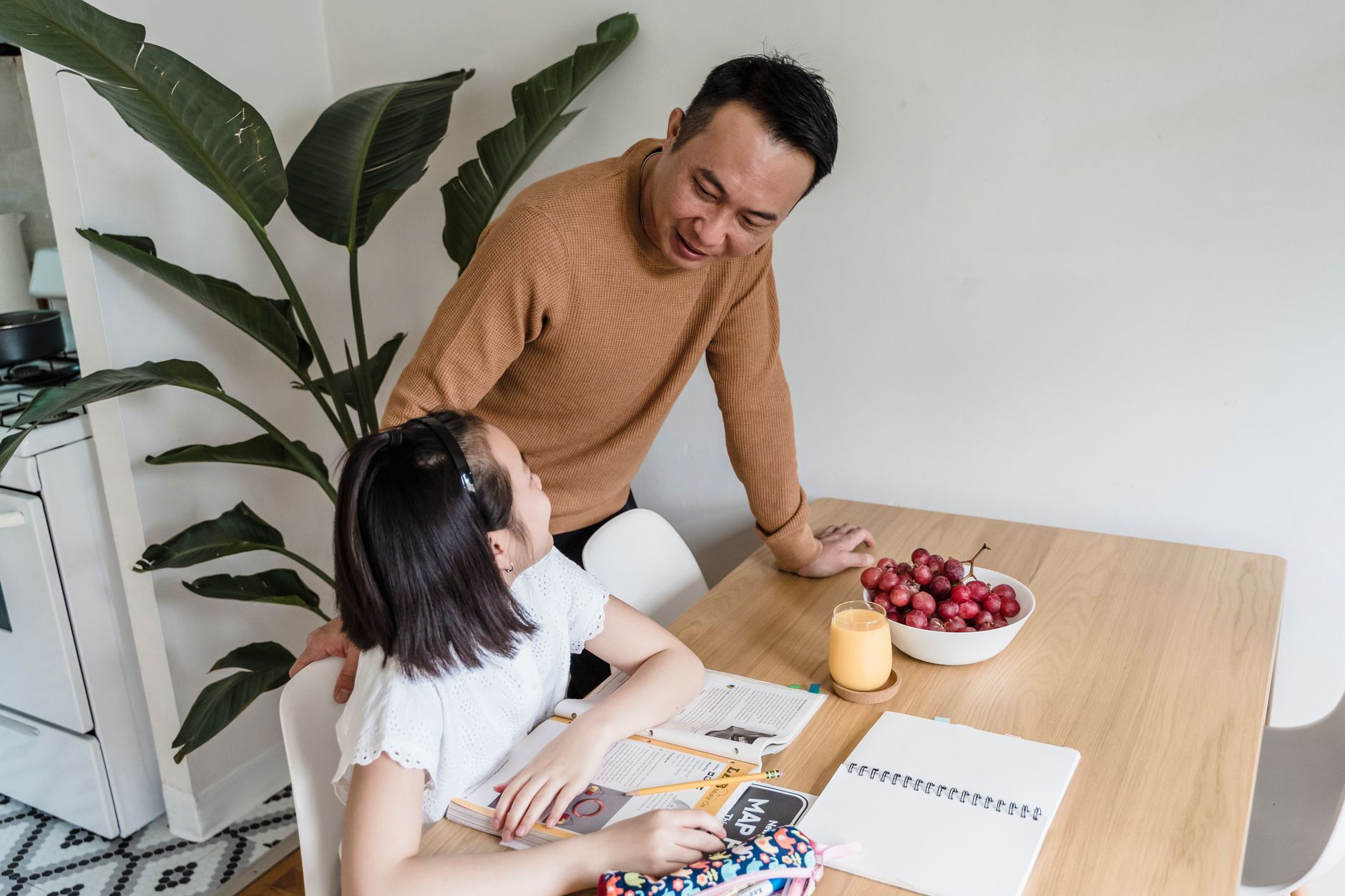 A father leans on the kitchen table and smiles at his daughter as she does homework with a textbook and notebook