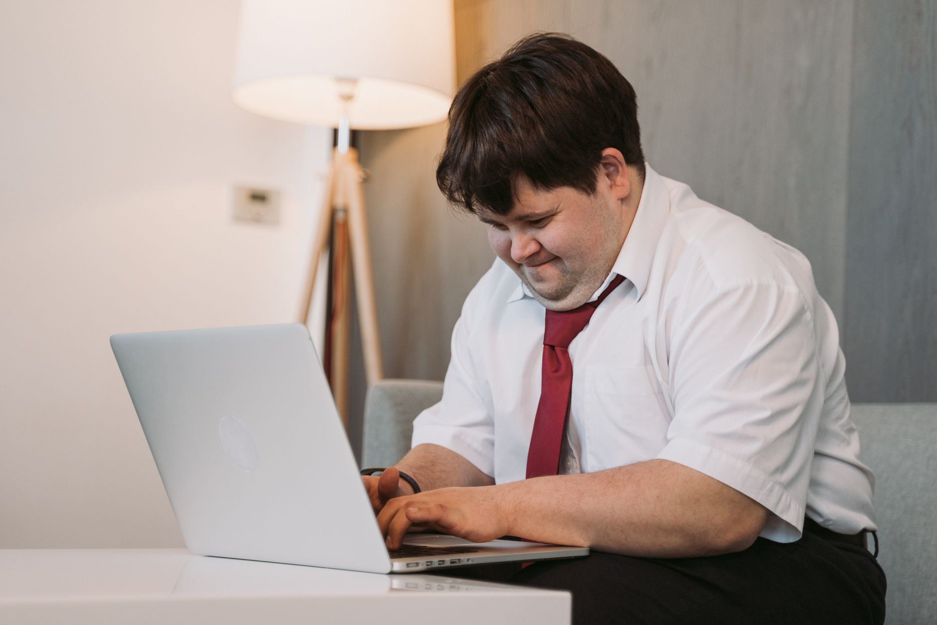 A smiling young man with Down syndrome in a white shirt and red tie typing on a laptop