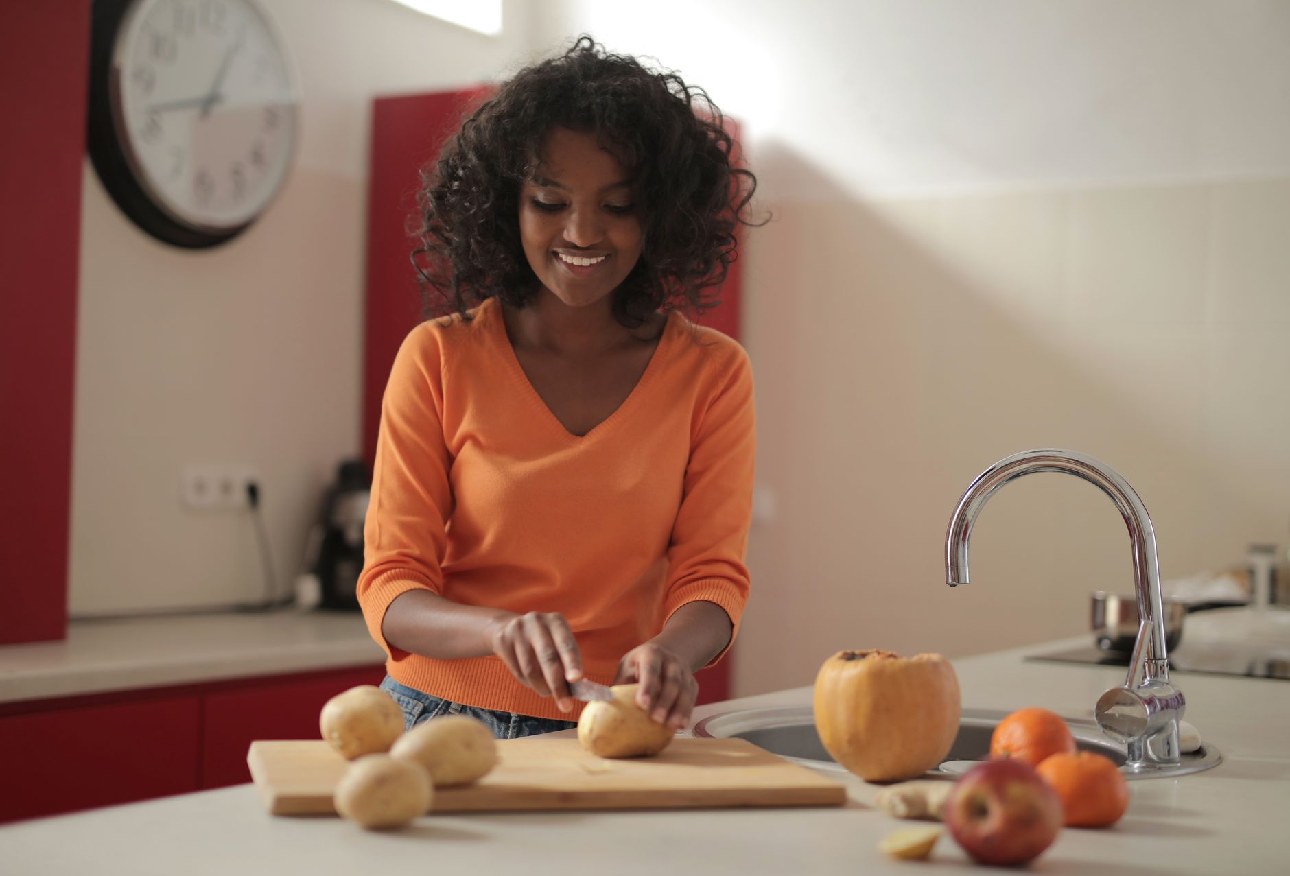 A smiling woman in an orange sweater slices potatoes on a cutting board in her kitchen