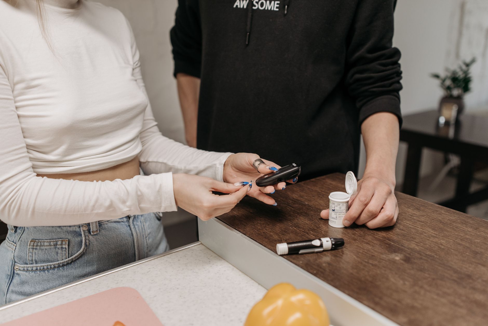 Two people at a kitchen counter, one holding a blood glucose meter and lancet while the other holds a vial of test strips