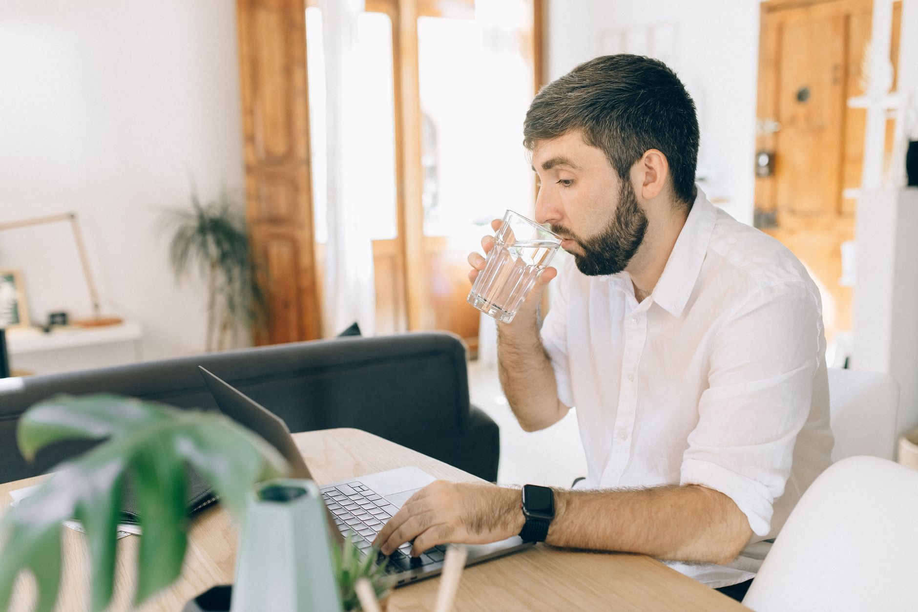 A bearded man in a white shirt drinks a glass of water while typing on a laptop at home