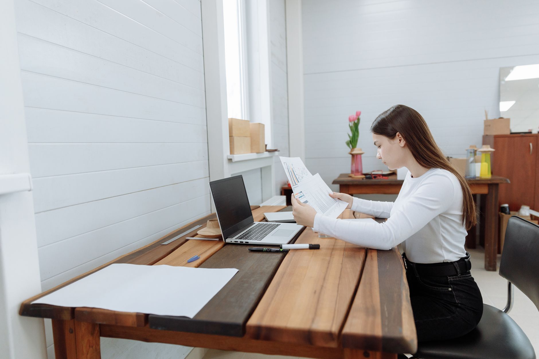 A young woman in a white sweater sits at a long wooden table reading printed charts beside her laptop