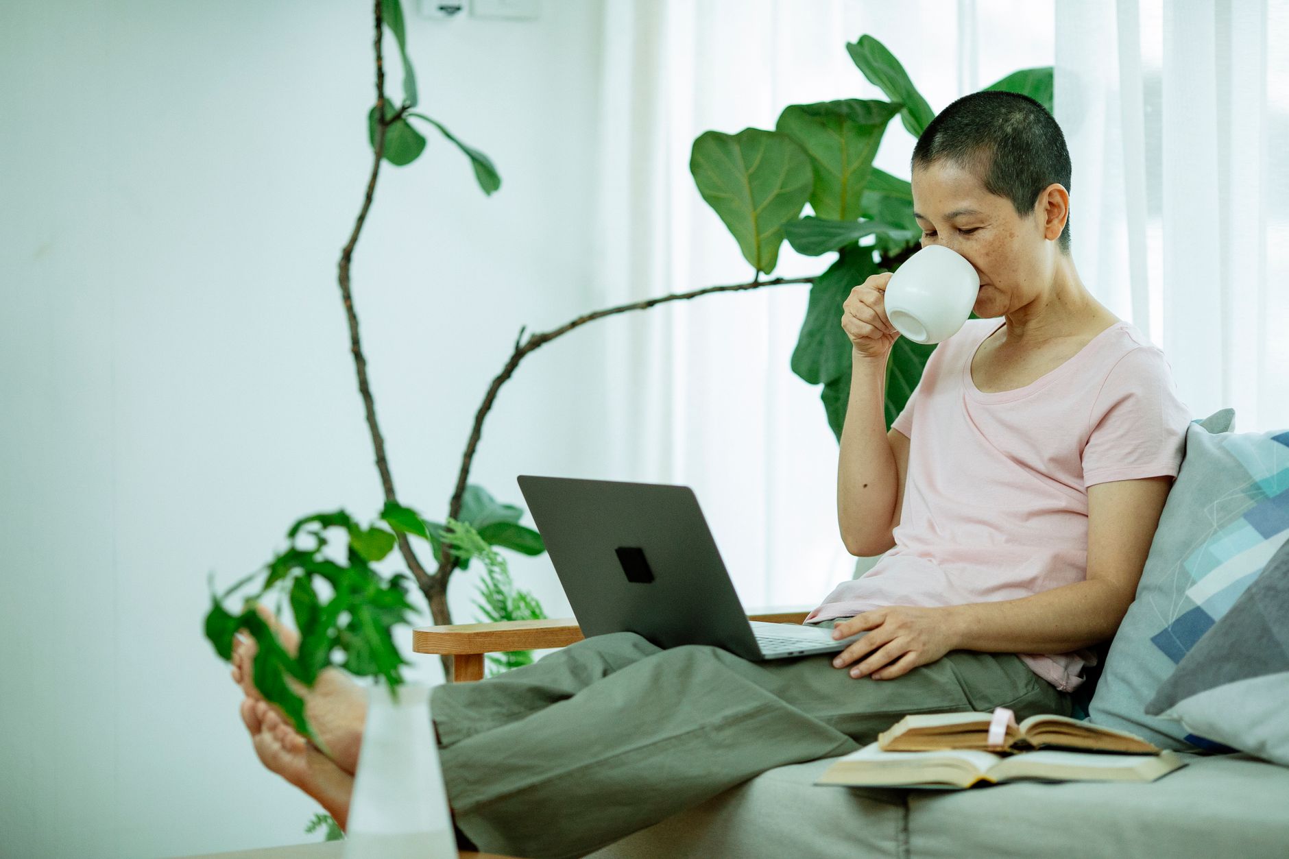 A woman with short-cropped hair sips from a mug while working on a laptop on her sofa at home
