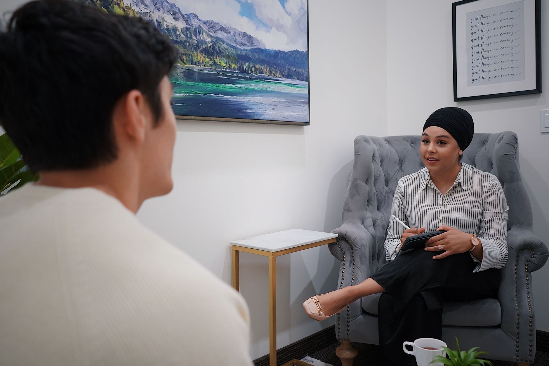 A counselor in a striped shirt and dark turban takes notes on a tablet while talking with a young man in a calm office