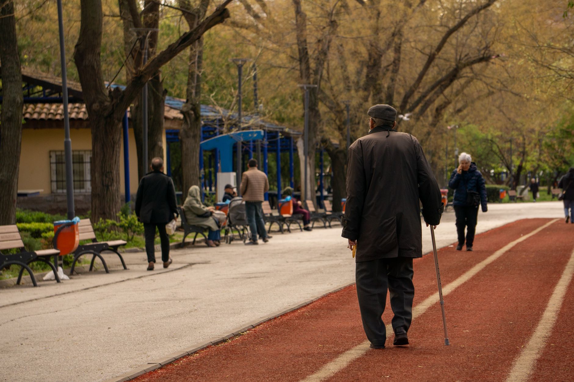 An older man in a coat and cap walks with a cane along a red path in a city park
