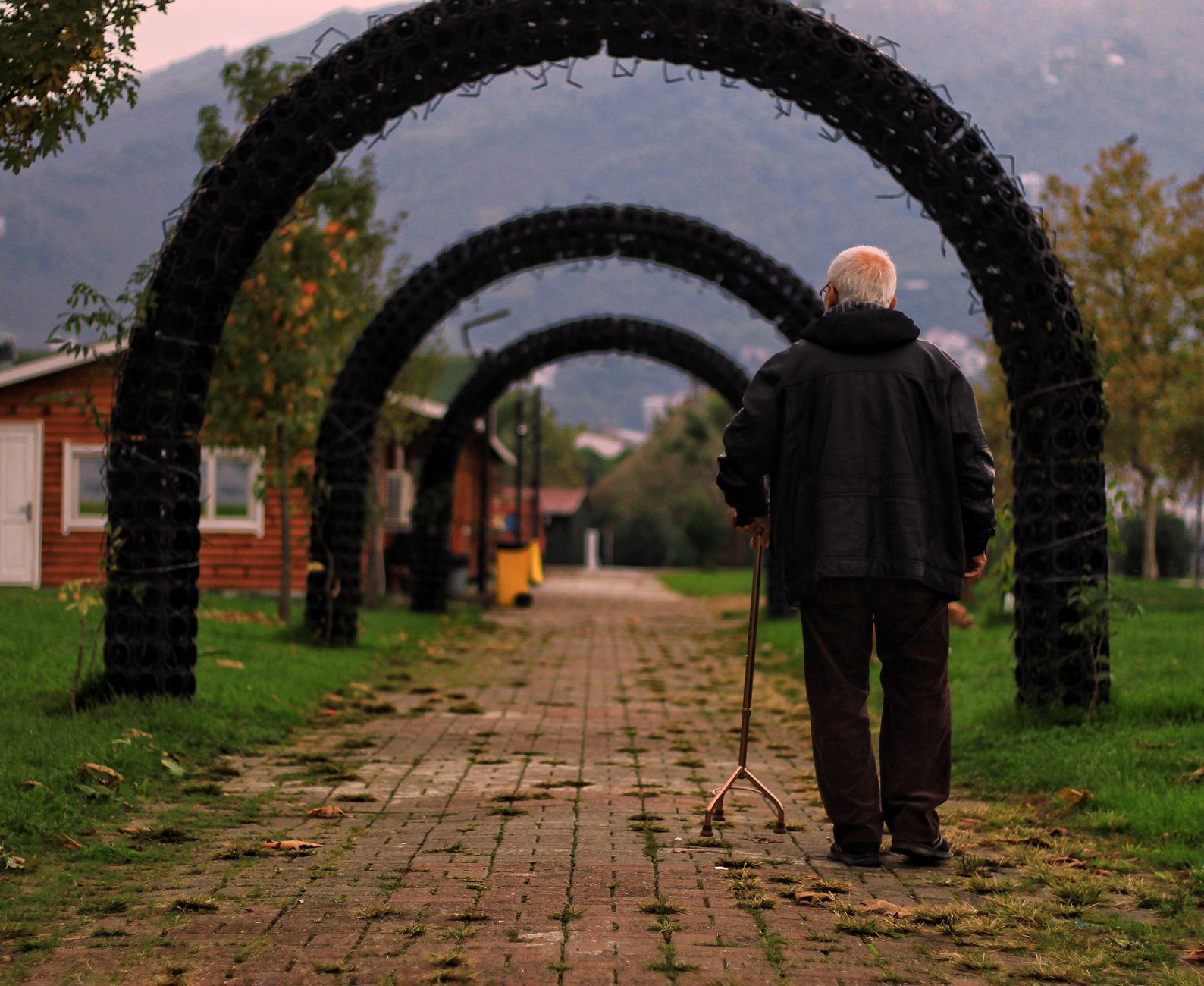 An older man using a quad cane walks along a brick path under a row of arches in a park