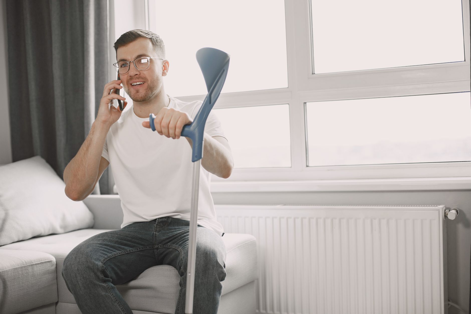A man holding a forearm crutch talks on his phone while sitting on a couch by a window