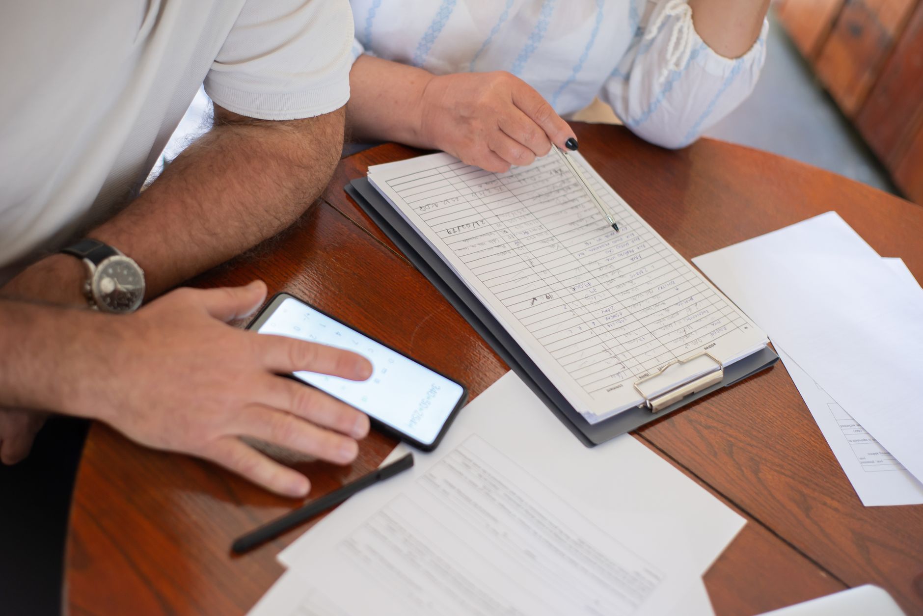 Two people at a wooden table go over handwritten figures on a clipboard while one uses a phone calculator