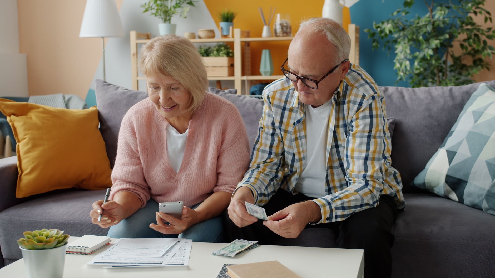 An older couple on a sofa go over household paperwork together with a phone and pen
