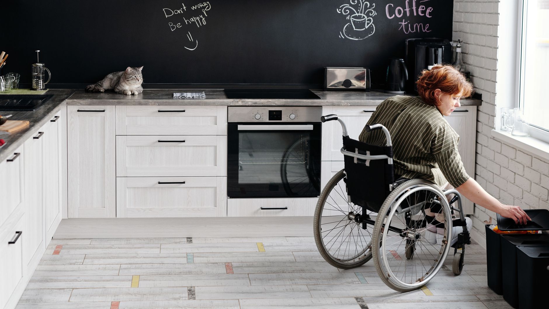 A woman using a wheelchair reaches into a low cabinet in her bright kitchen while a tabby cat rests on the counter