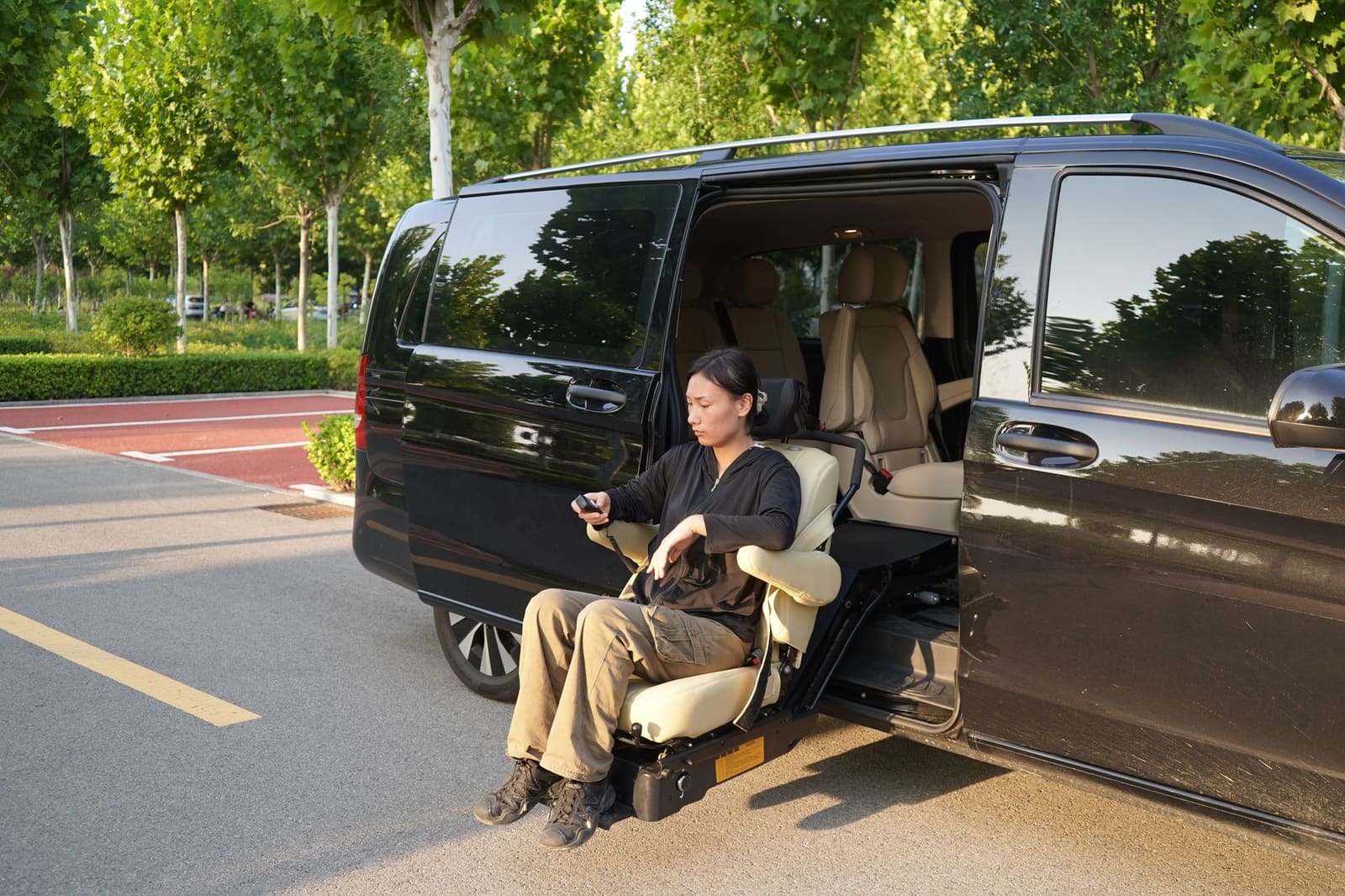 A woman uses a powered swivel seat to lower herself out of the side door of a black van in a tree-lined parking lot