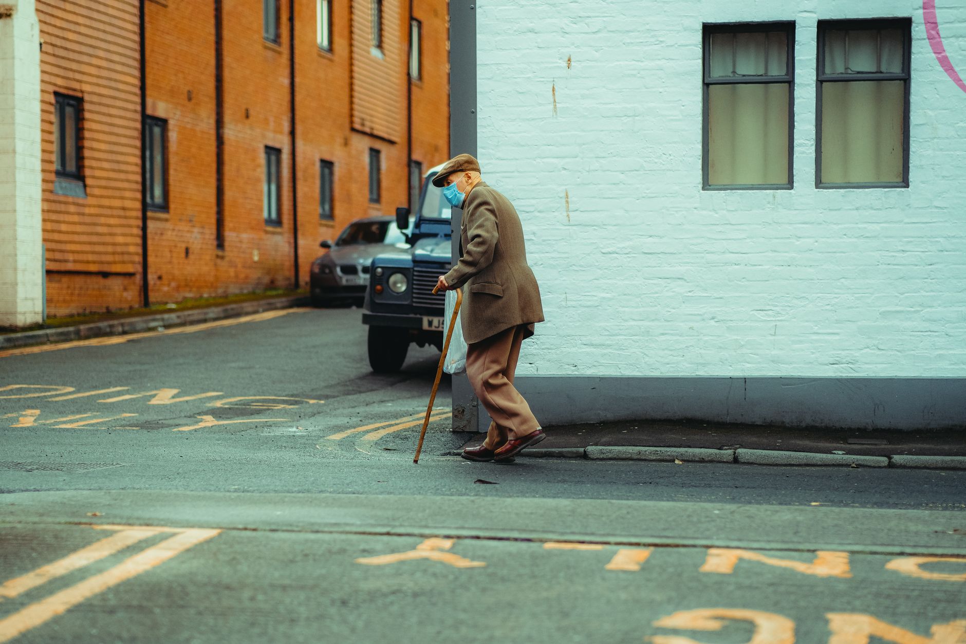 An older man in a flat cap walks with a wooden cane past a white brick building