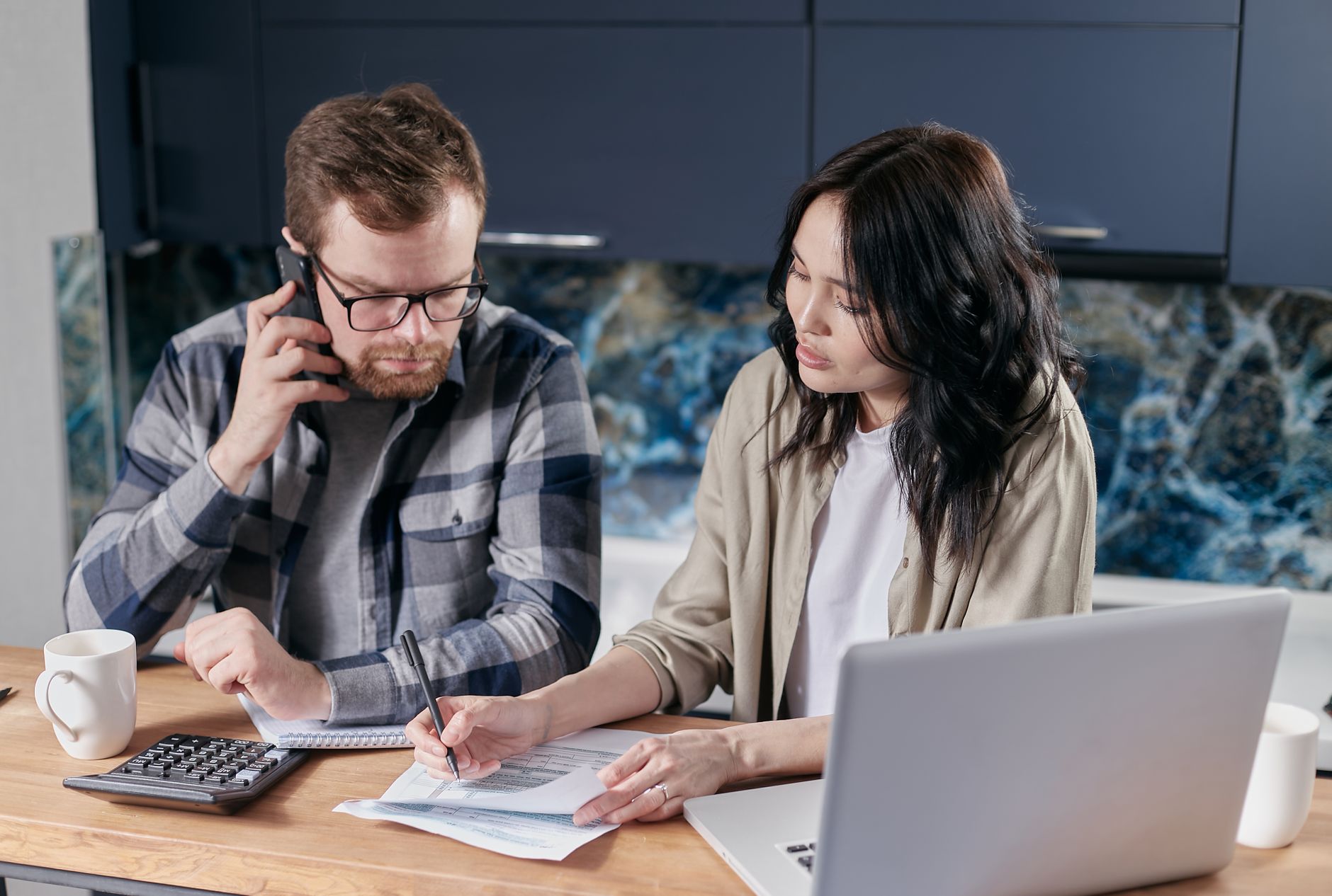 A man on the phone and a woman filling out a form sit together at a kitchen table with a laptop and calculator