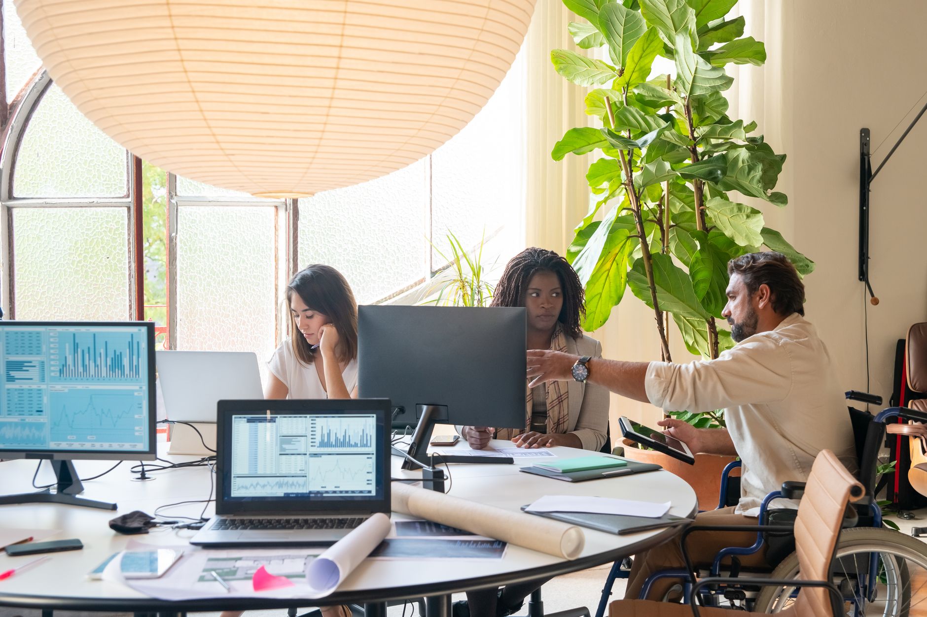 A man using a wheelchair points across a shared office desk while talking with two coworkers at their computers