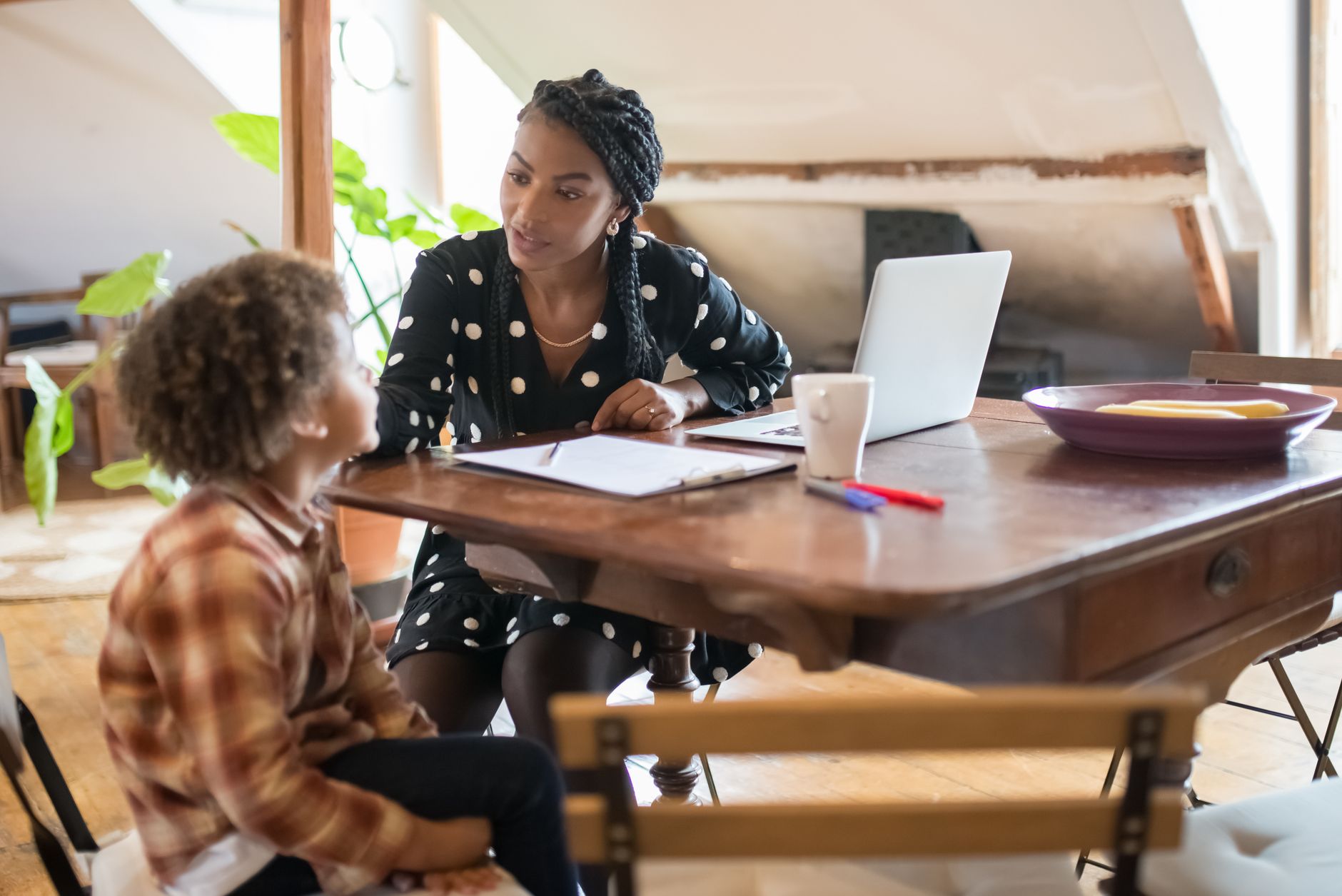 A mother sits at a wooden dining table with a laptop and clipboard, listening closely to her young son