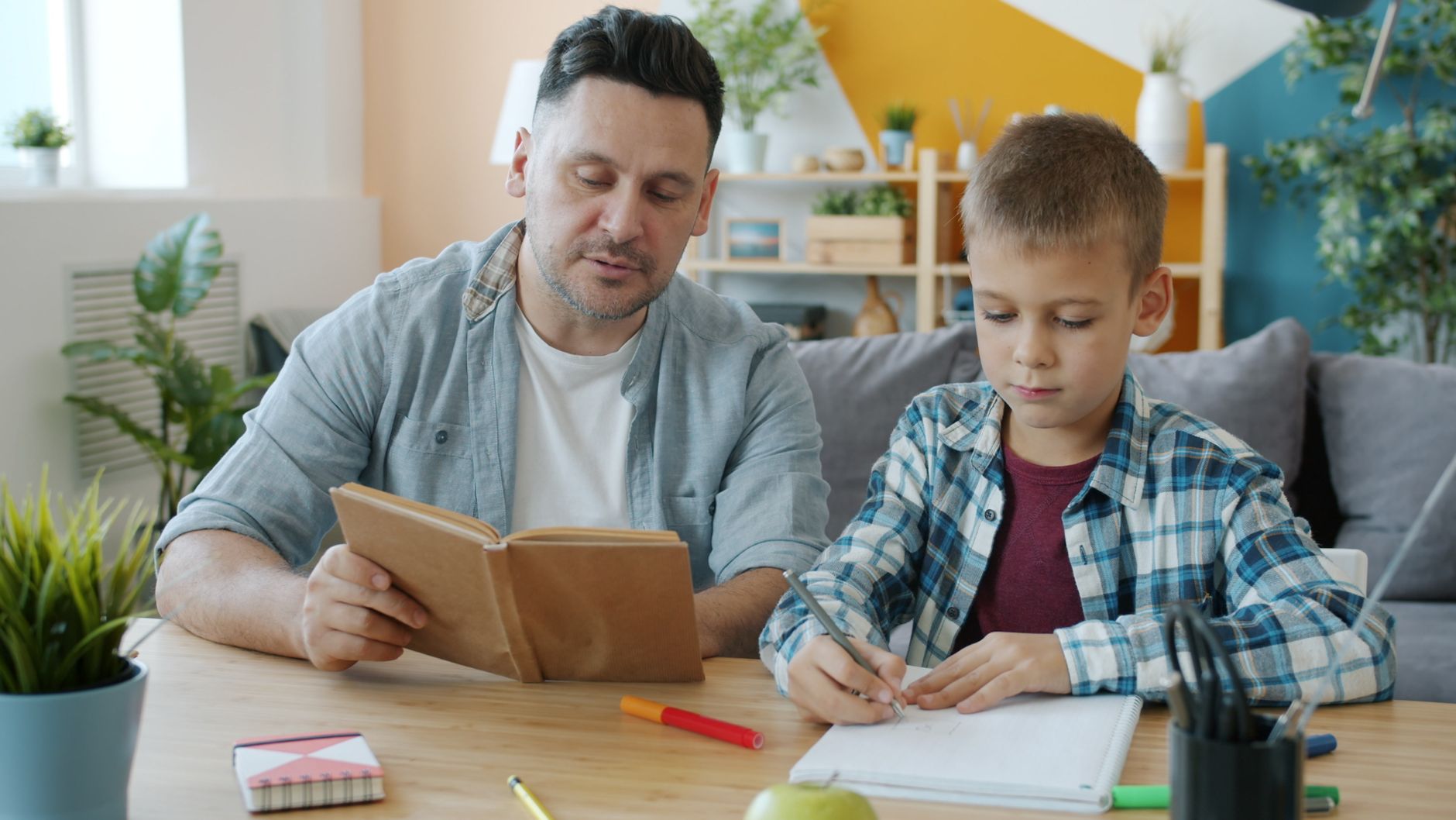 A father reads from a book while his young son writes in a notebook at a wooden table at home