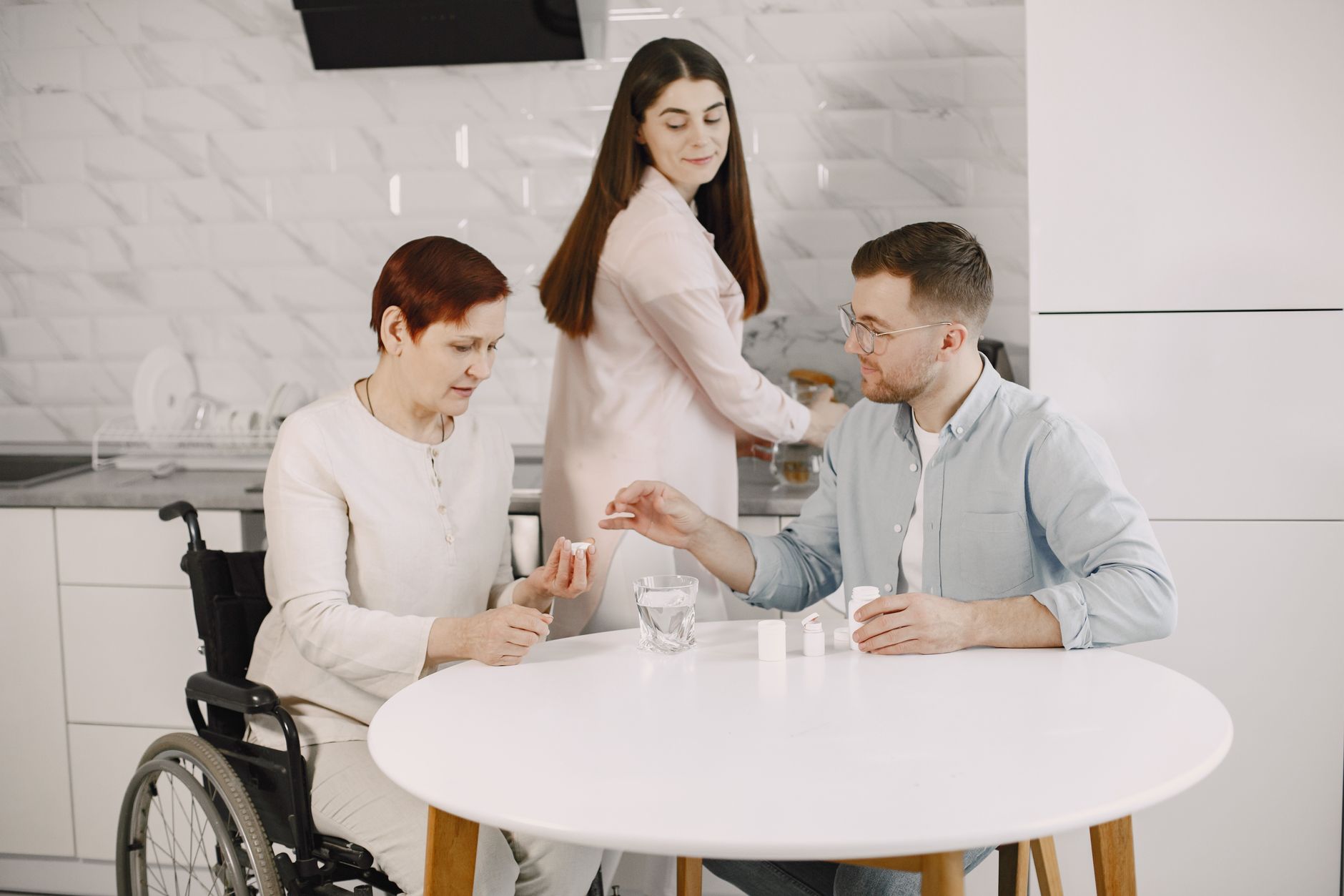 An older woman who uses a wheelchair sorts her medication at a kitchen table while her adult son hands her a pill and her daughter works at the counter