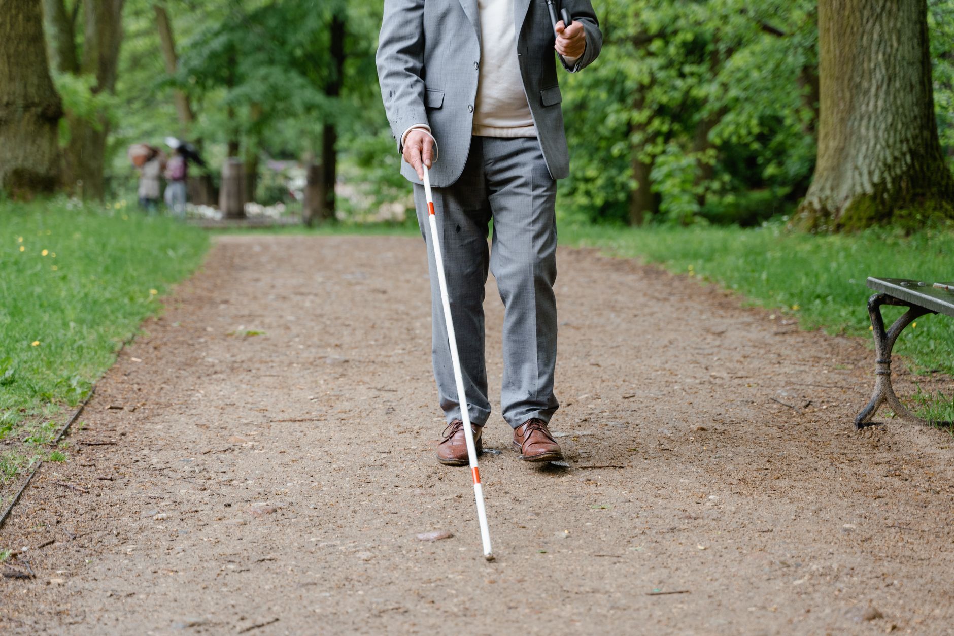 A man in a gray suit walks along a tree-lined park path using a white cane