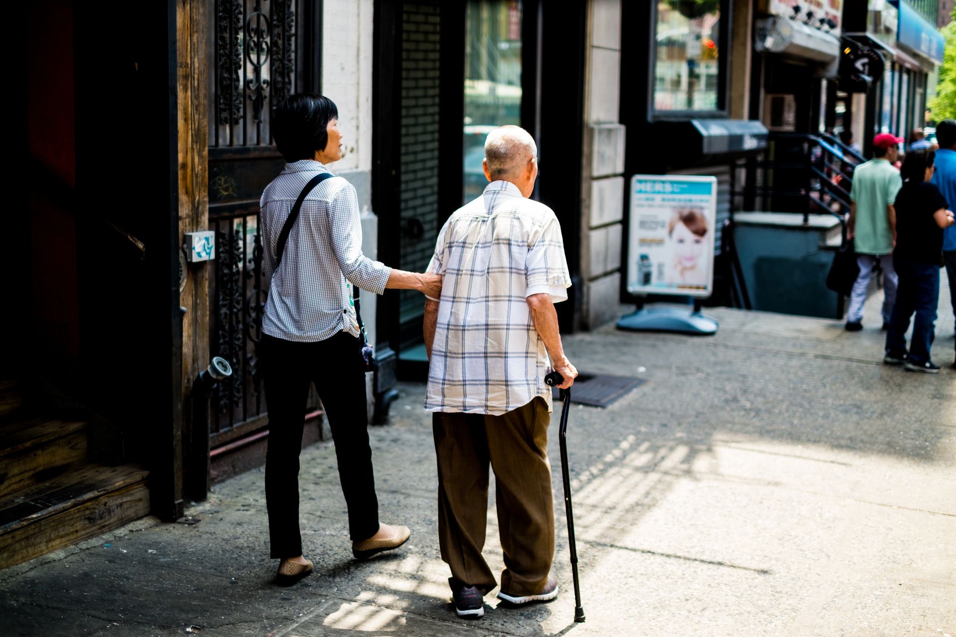 An older man walking with a cane on a busy New York sidewalk, with a woman beside him holding his arm