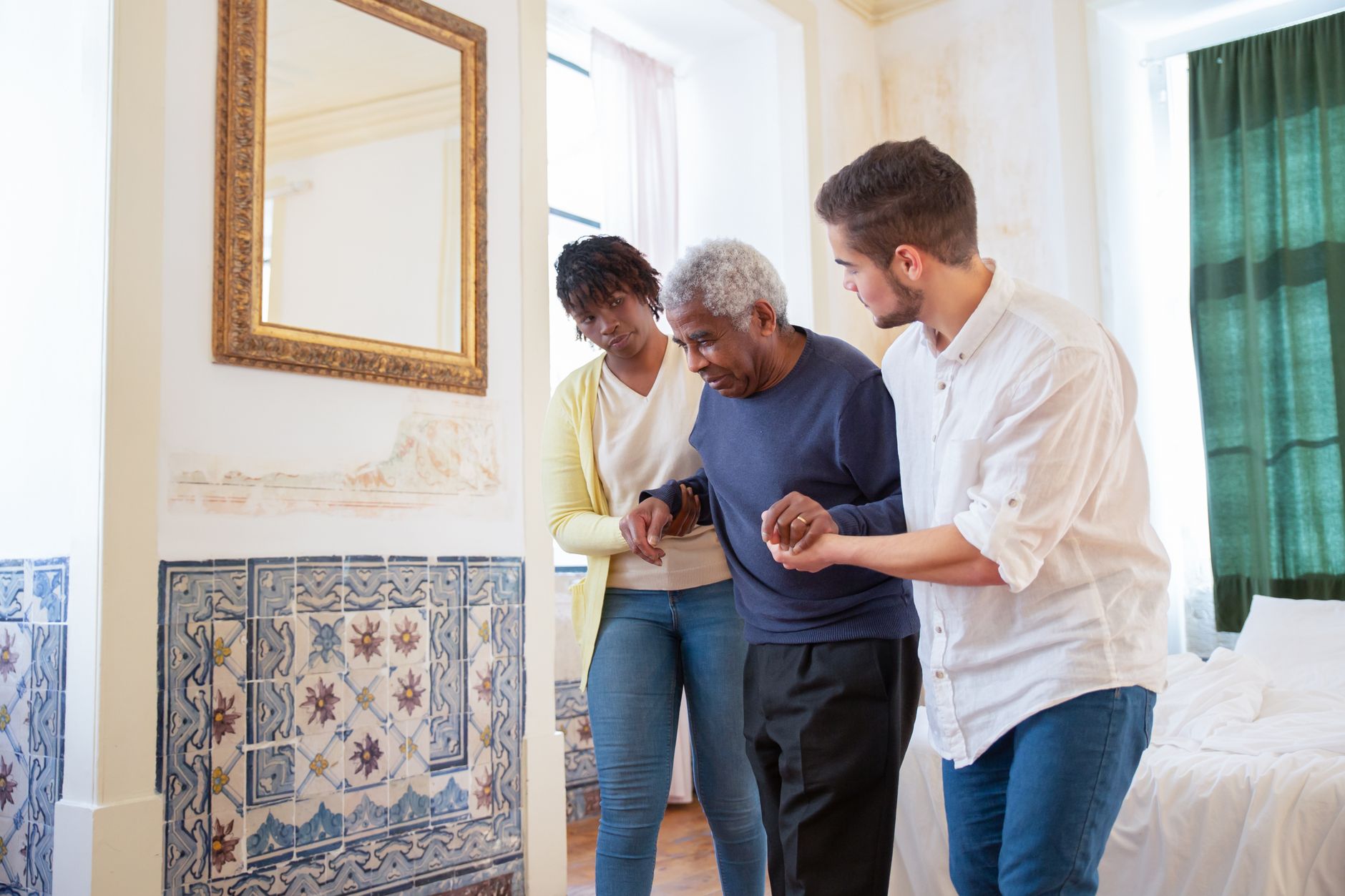 A young man and a woman support an older man by the arms as he walks across a sunny bedroom