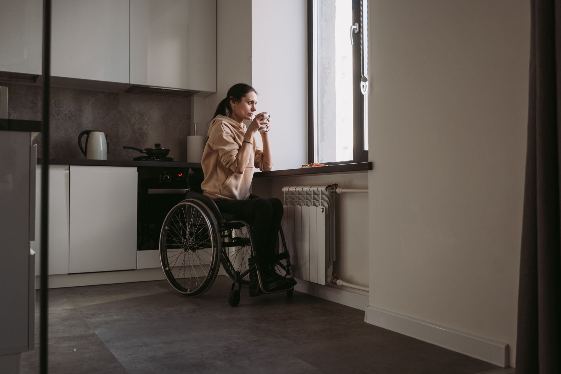 A woman using a wheelchair sips from a mug by the window of her bright apartment kitchen