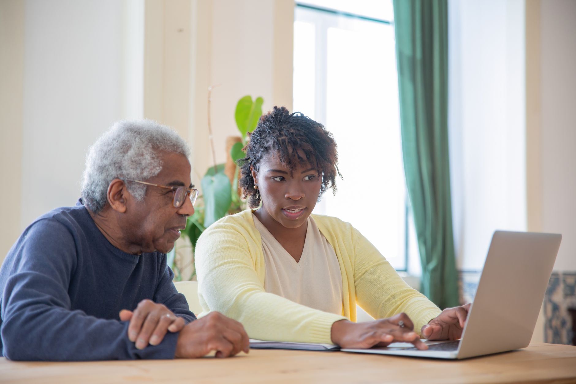 A young woman helps an older man compare information on a laptop at a dining table
