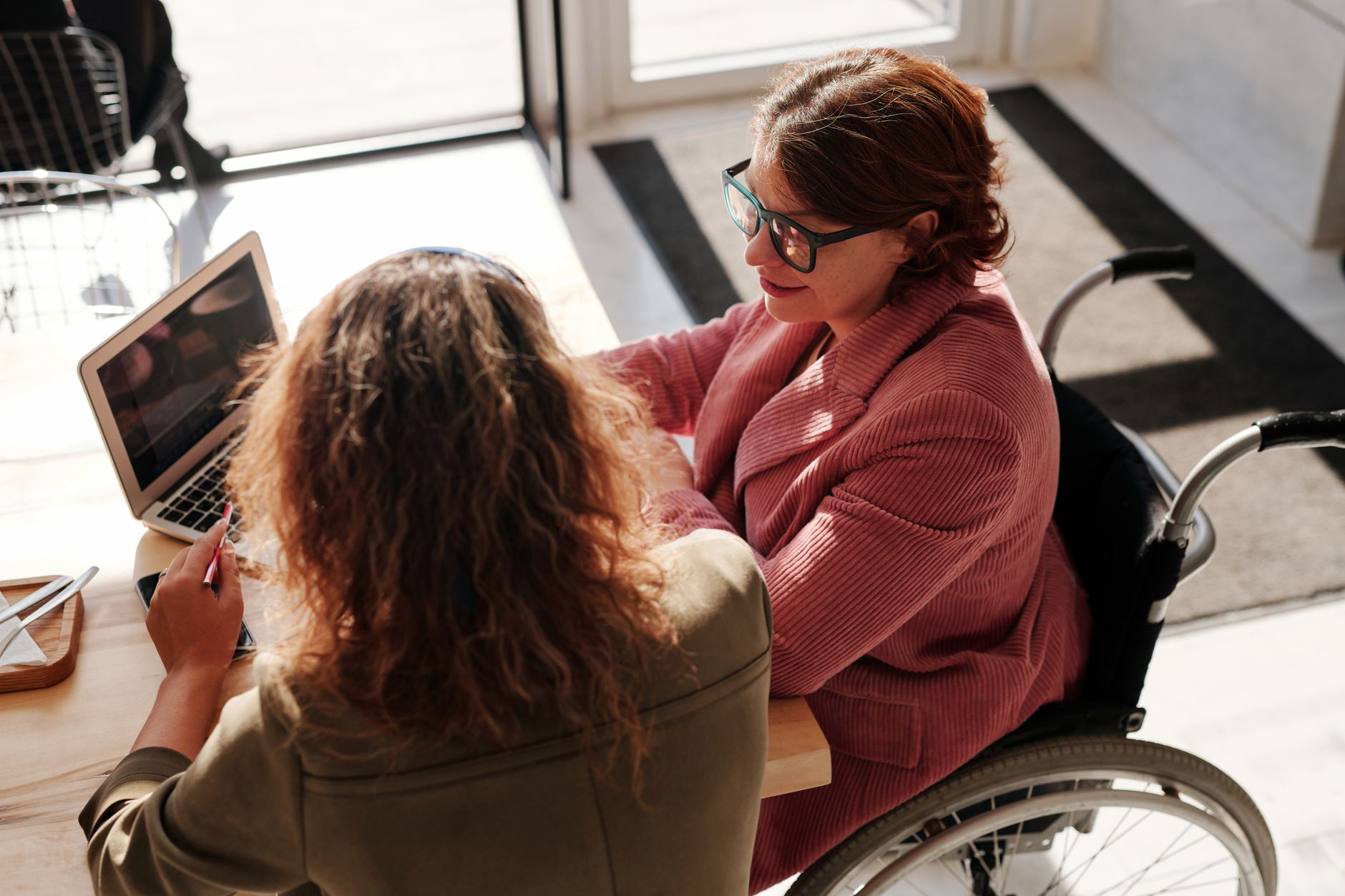 A woman using a wheelchair talks with a colleague over a laptop at a sunny cafe table