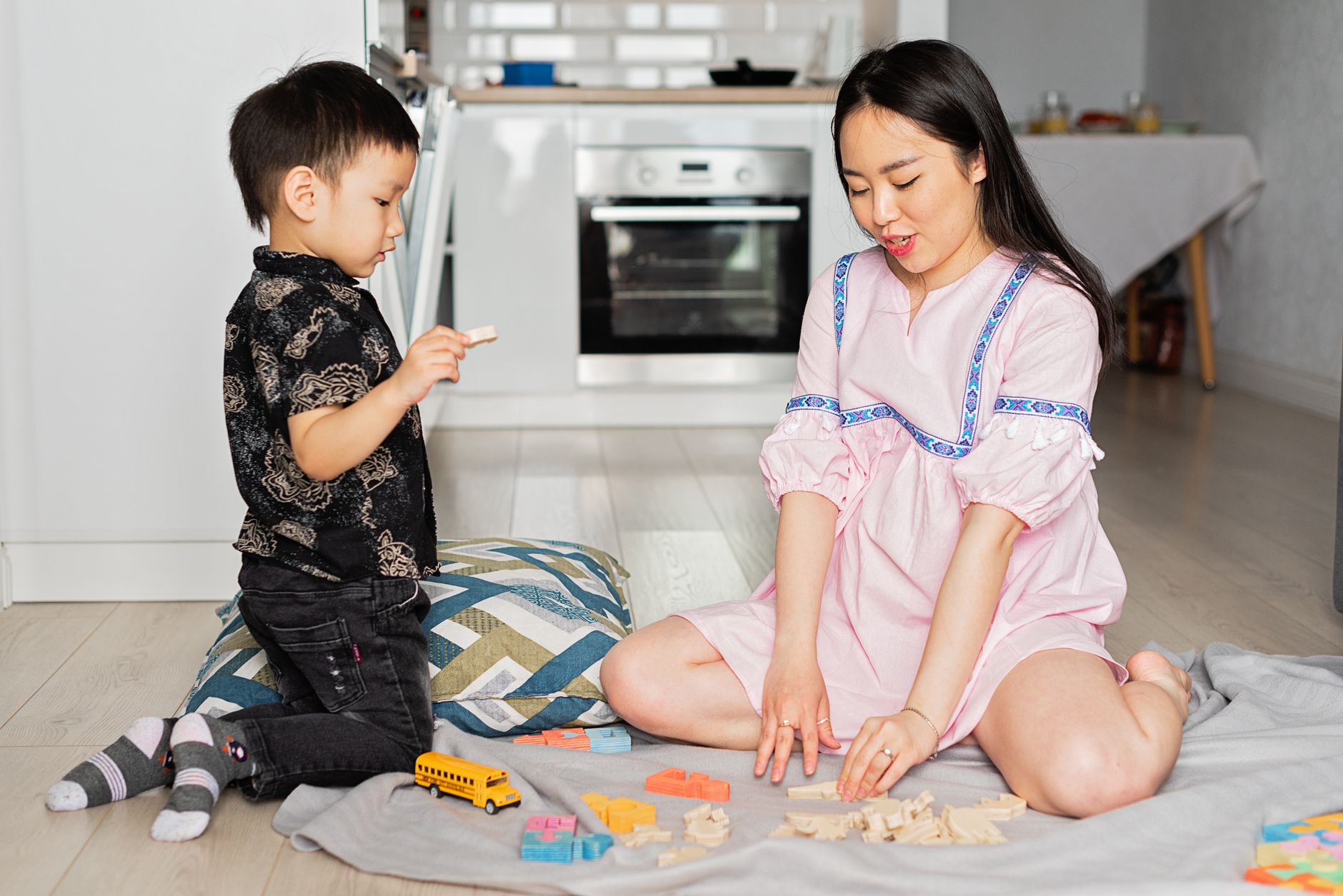 A young boy kneels on a floor cushion holding a wooden puzzle piece while his mother, sitting beside him on a blanket, sorts colorful shapes