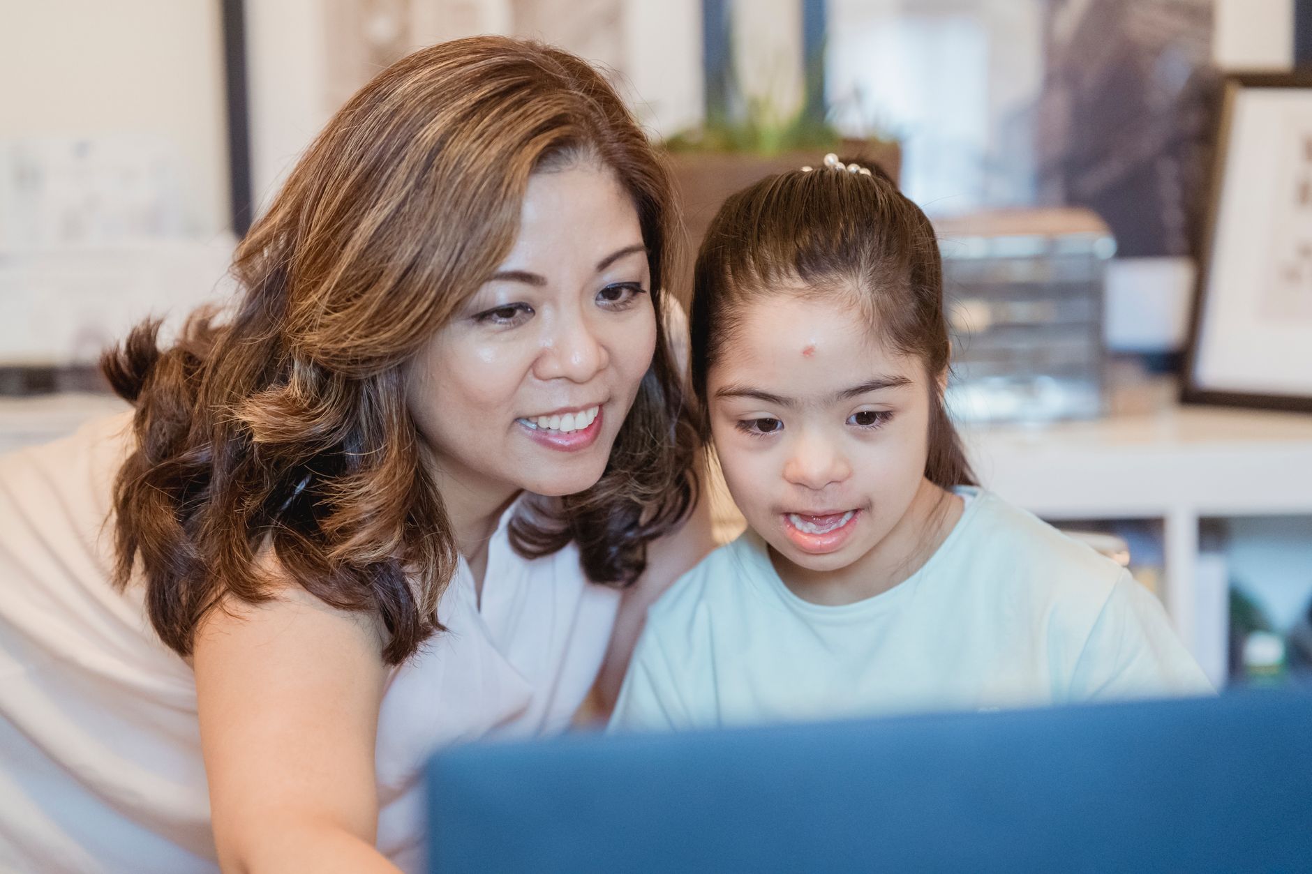 A smiling mother and her young daughter with Down syndrome look at a laptop together at home