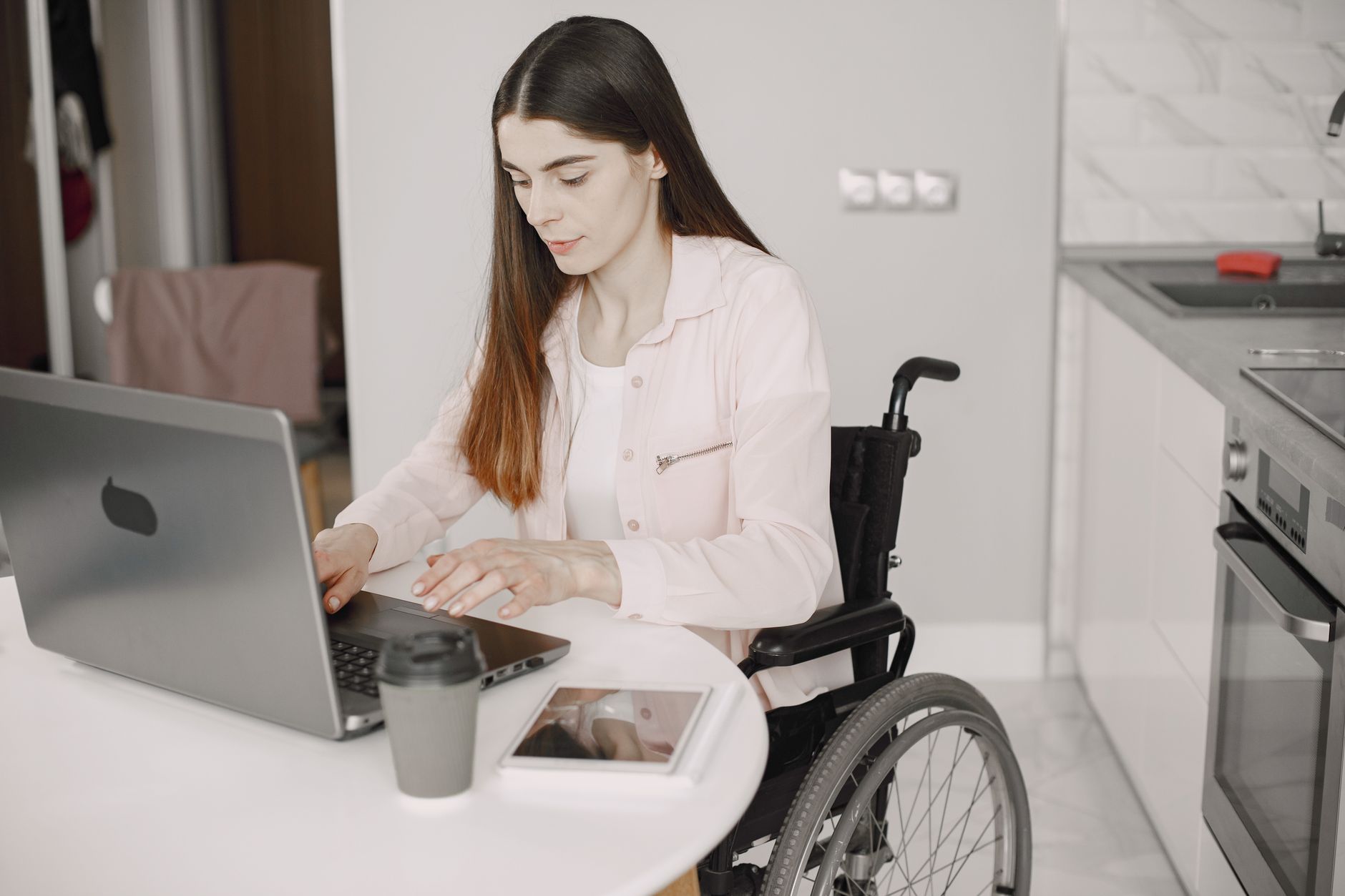 A woman using a wheelchair types on a laptop at a white table in her kitchen, with a coffee cup and tablet beside her