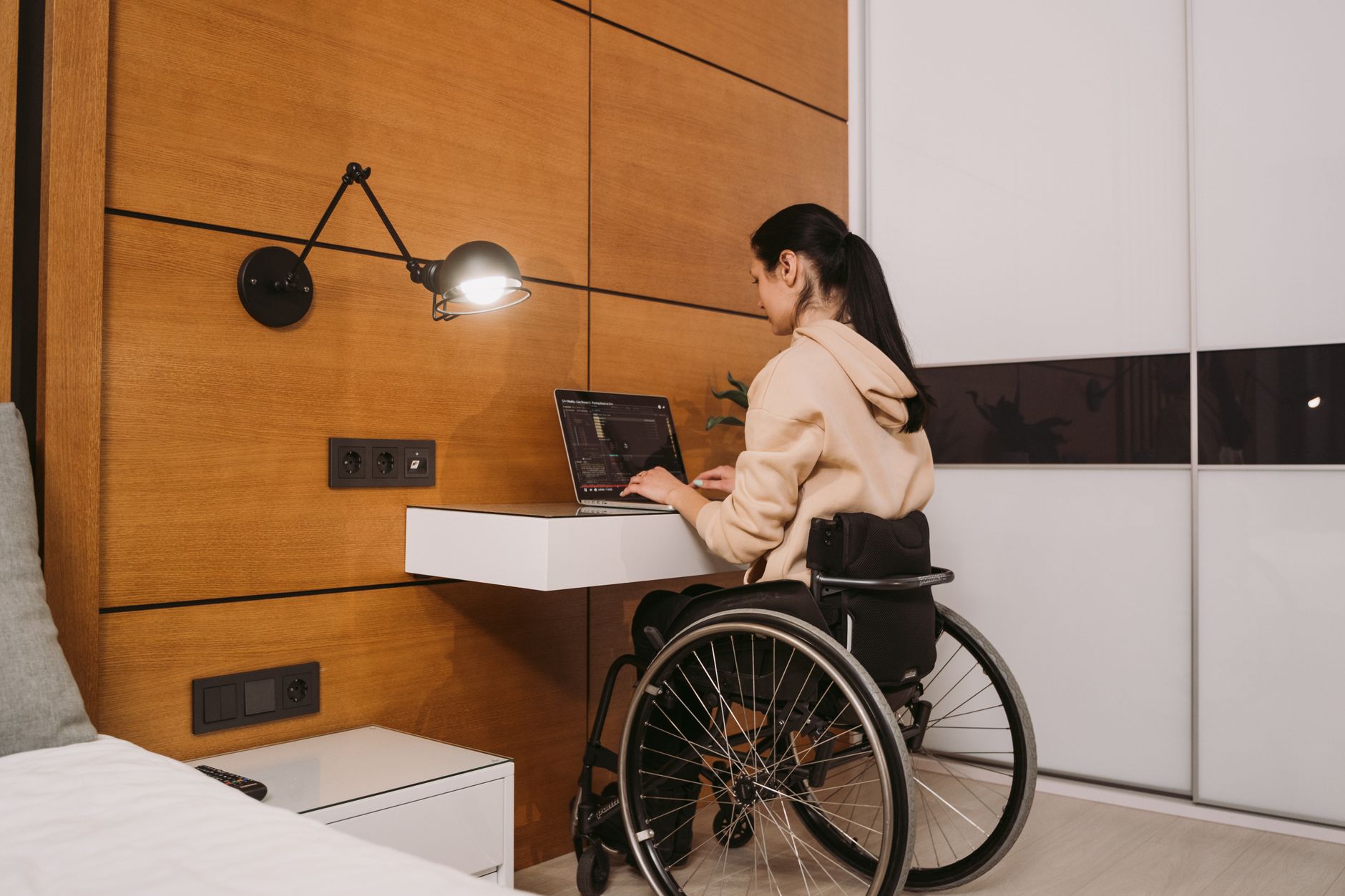 A woman using a wheelchair types on a laptop at a wall-mounted desk in her bedroom