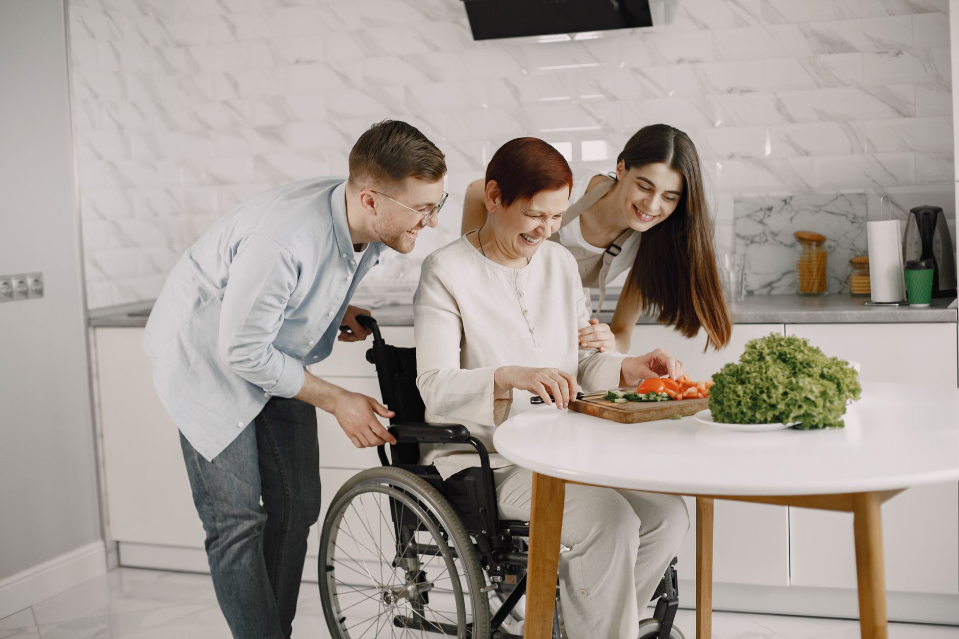 A woman using a wheelchair chops vegetables at a kitchen table while two younger family members lean in, laughing