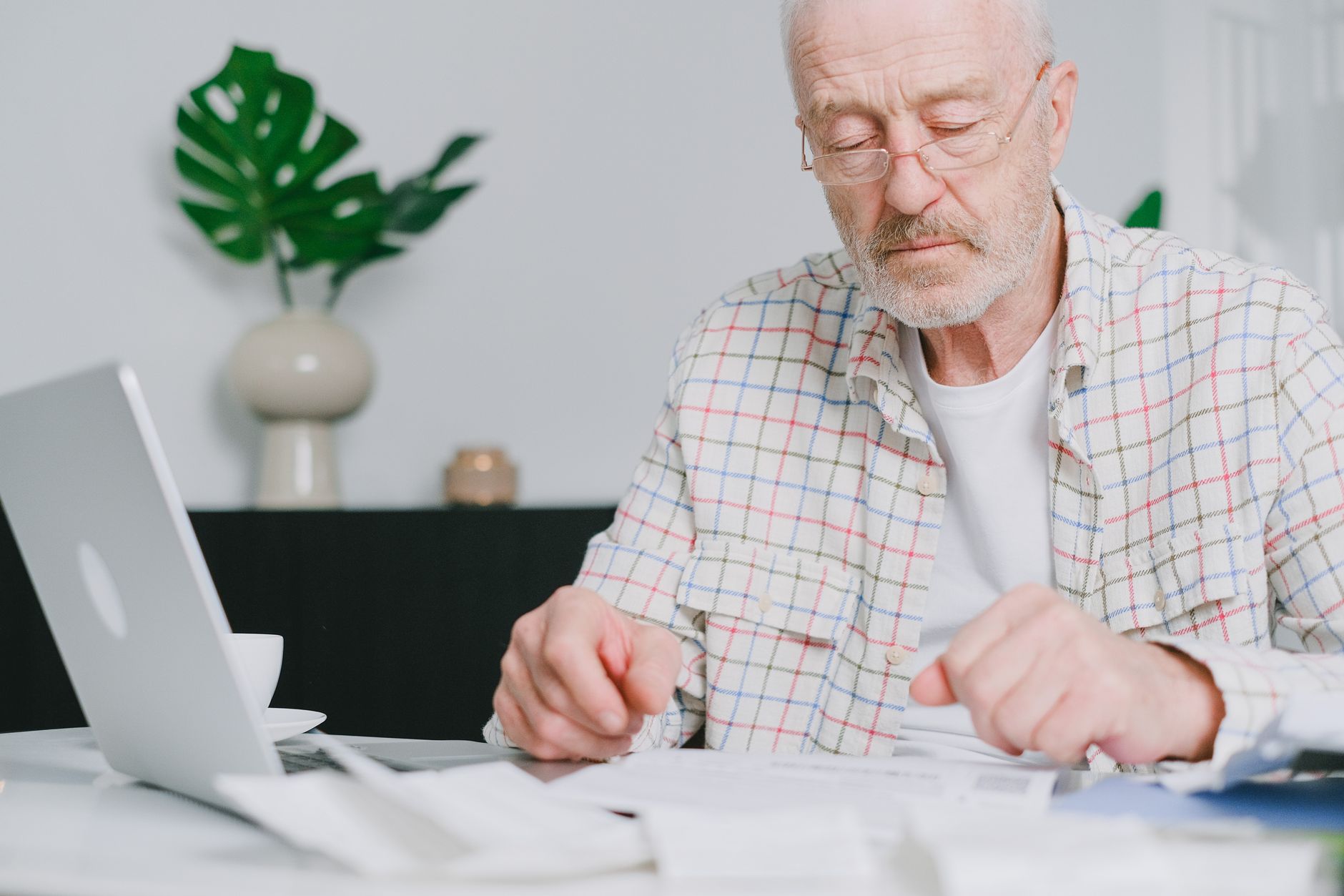 An older man in glasses and a plaid shirt reads through papers at a table beside a laptop