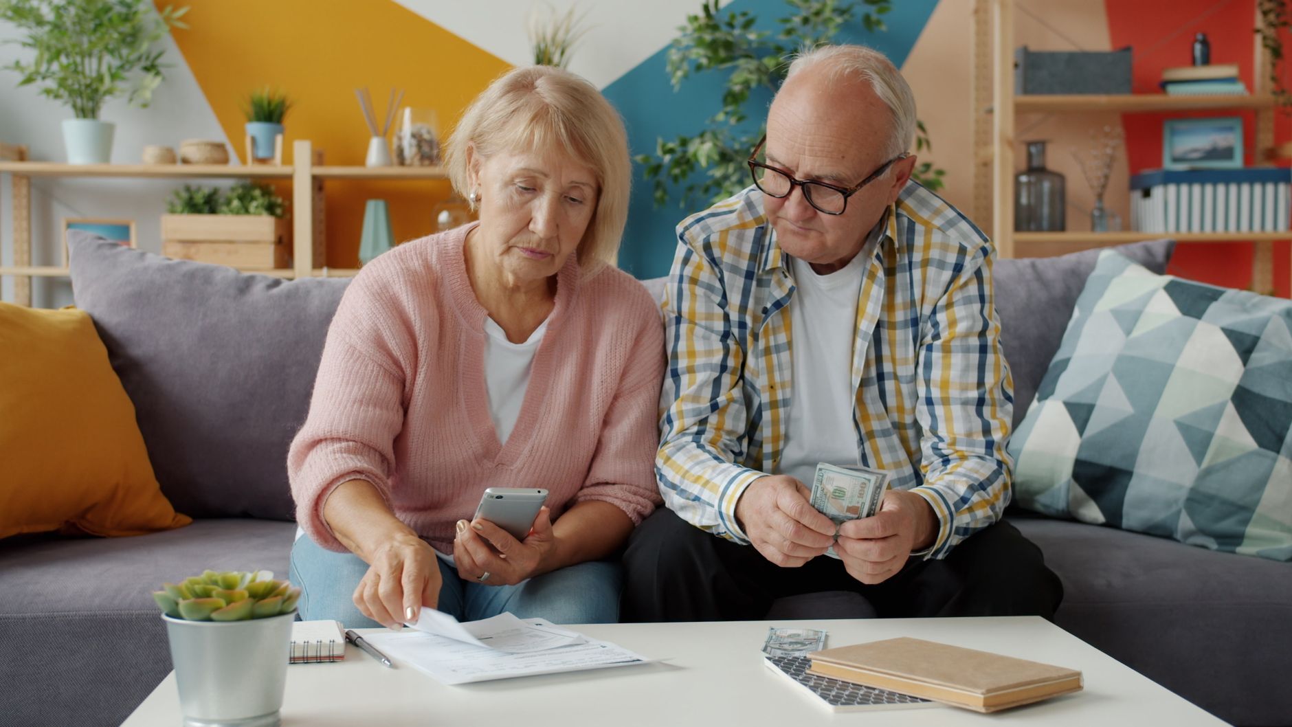 An older couple sits on a sofa going over household bills, one holding a phone and the other counting cash