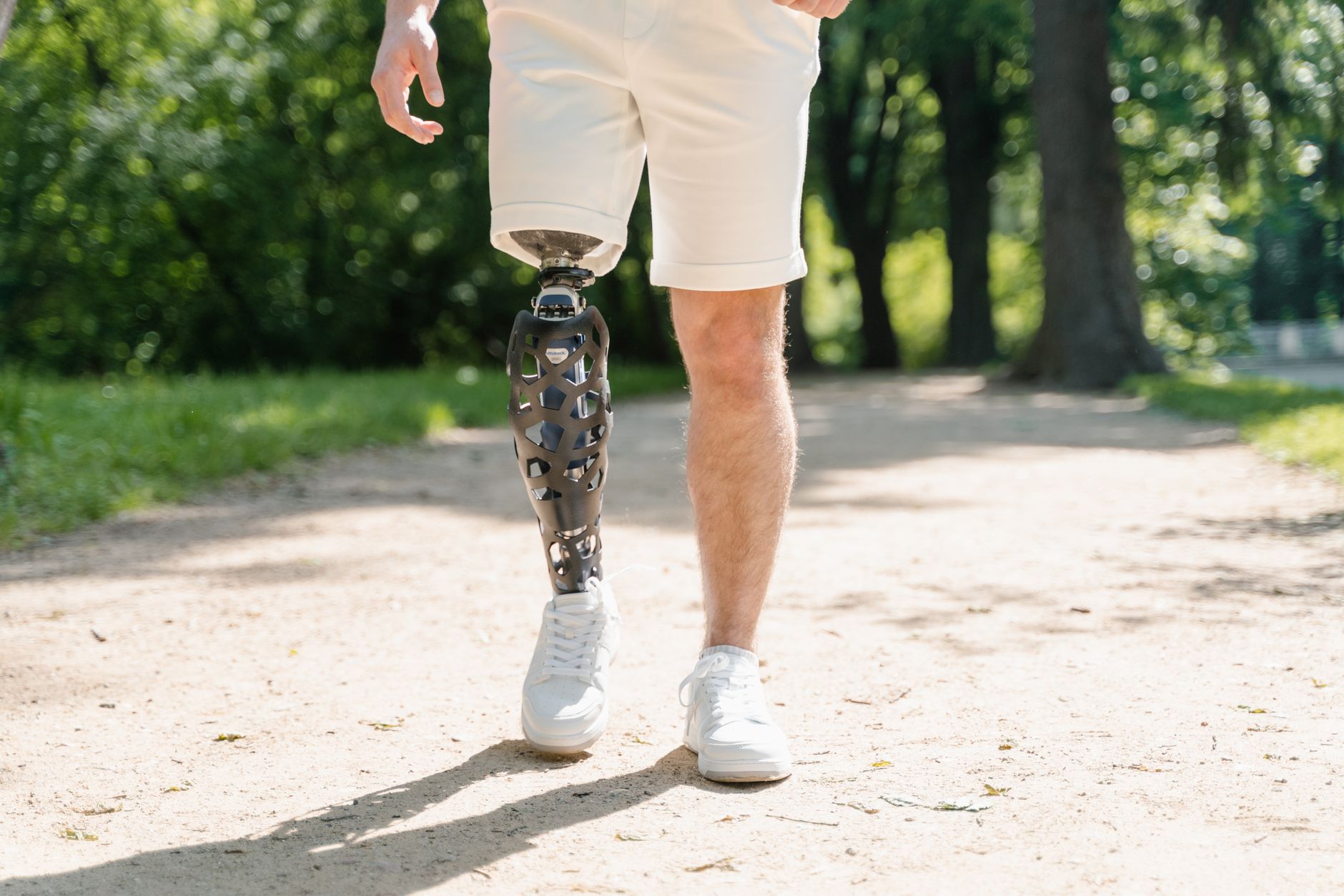 A man wearing a prosthetic leg and white sneakers walks along a sunny park path