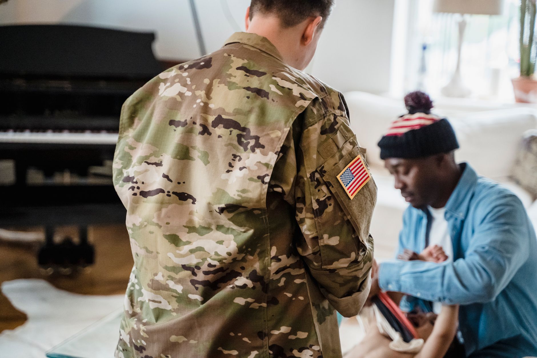 A service member in uniform stands in a living room while a family member kneels to dress a baby