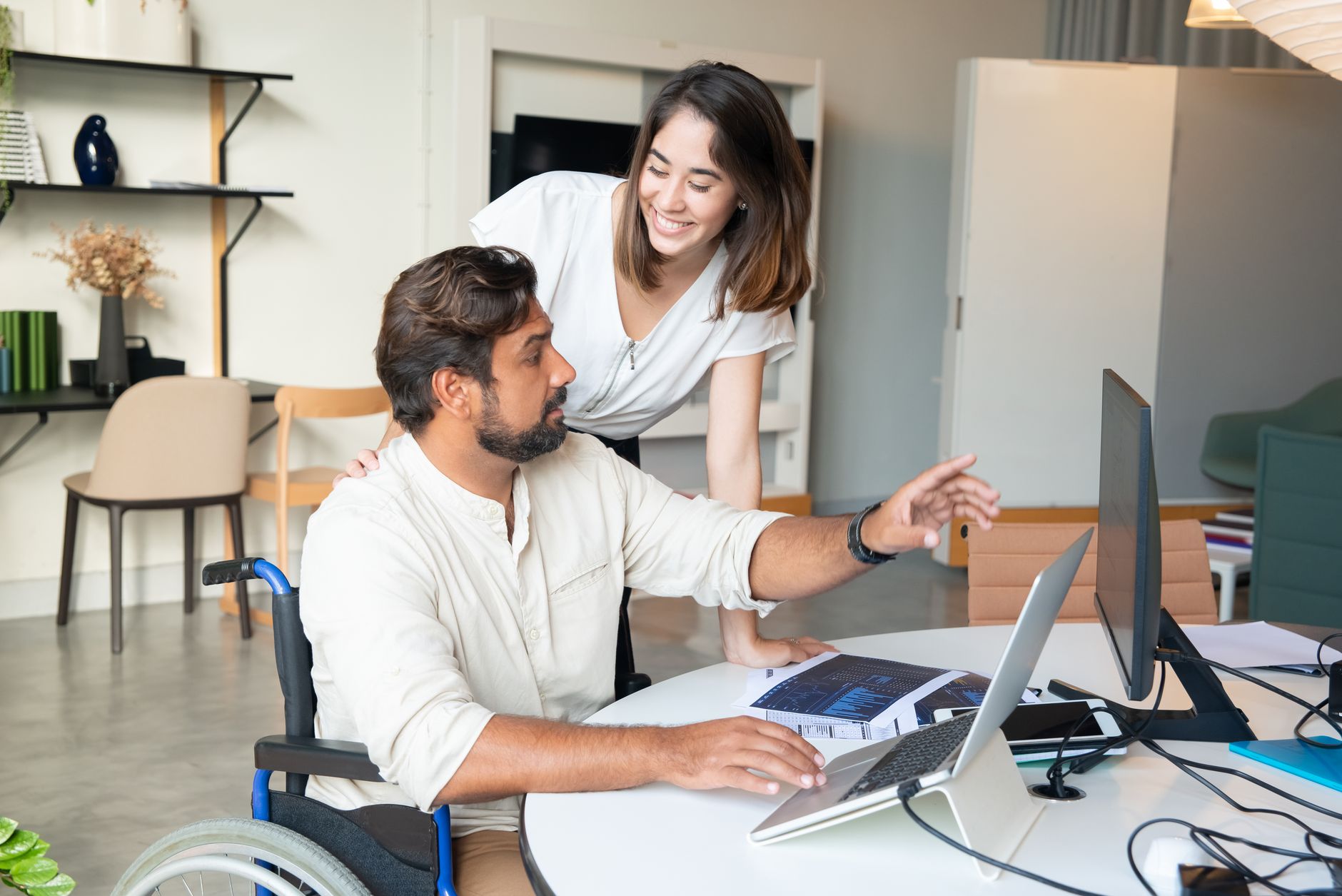 A man in a wheelchair points at his laptop screen at an office desk while a smiling coworker leans in beside him
