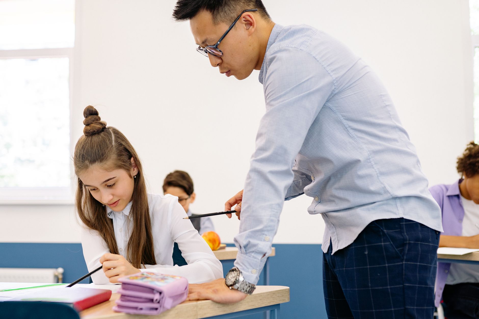 A teacher leans over a student's desk to point at her work with a pencil while classmates write in the background