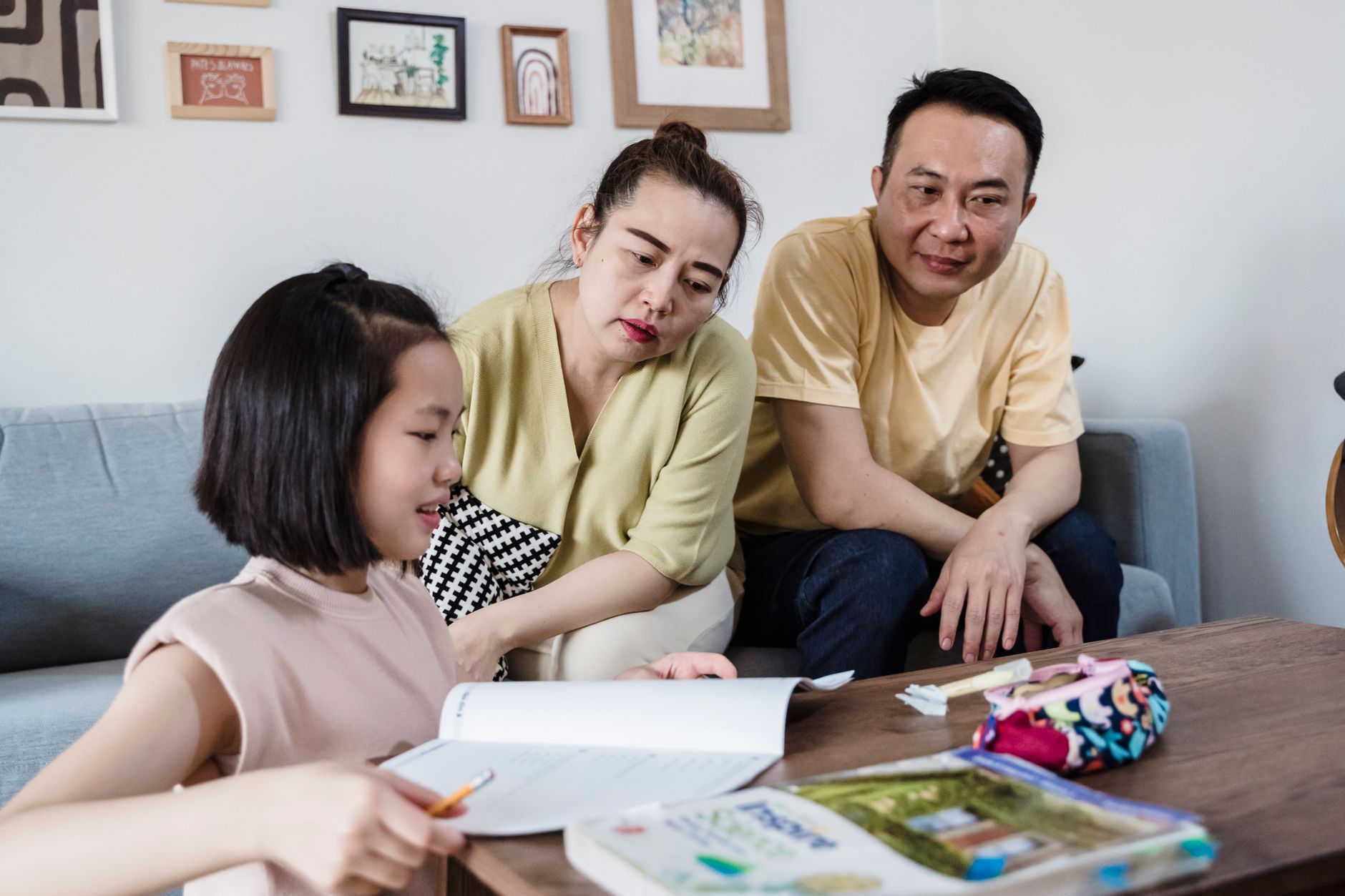 A girl works in a school workbook at a coffee table while her mother and father lean in beside her on the sofa