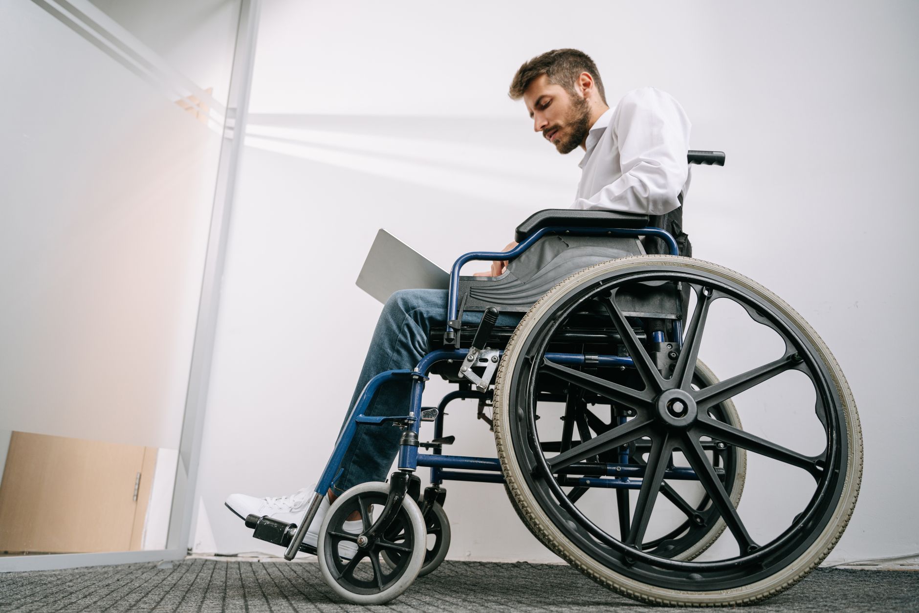 A man using a wheelchair works on a laptop in a bright office corridor
